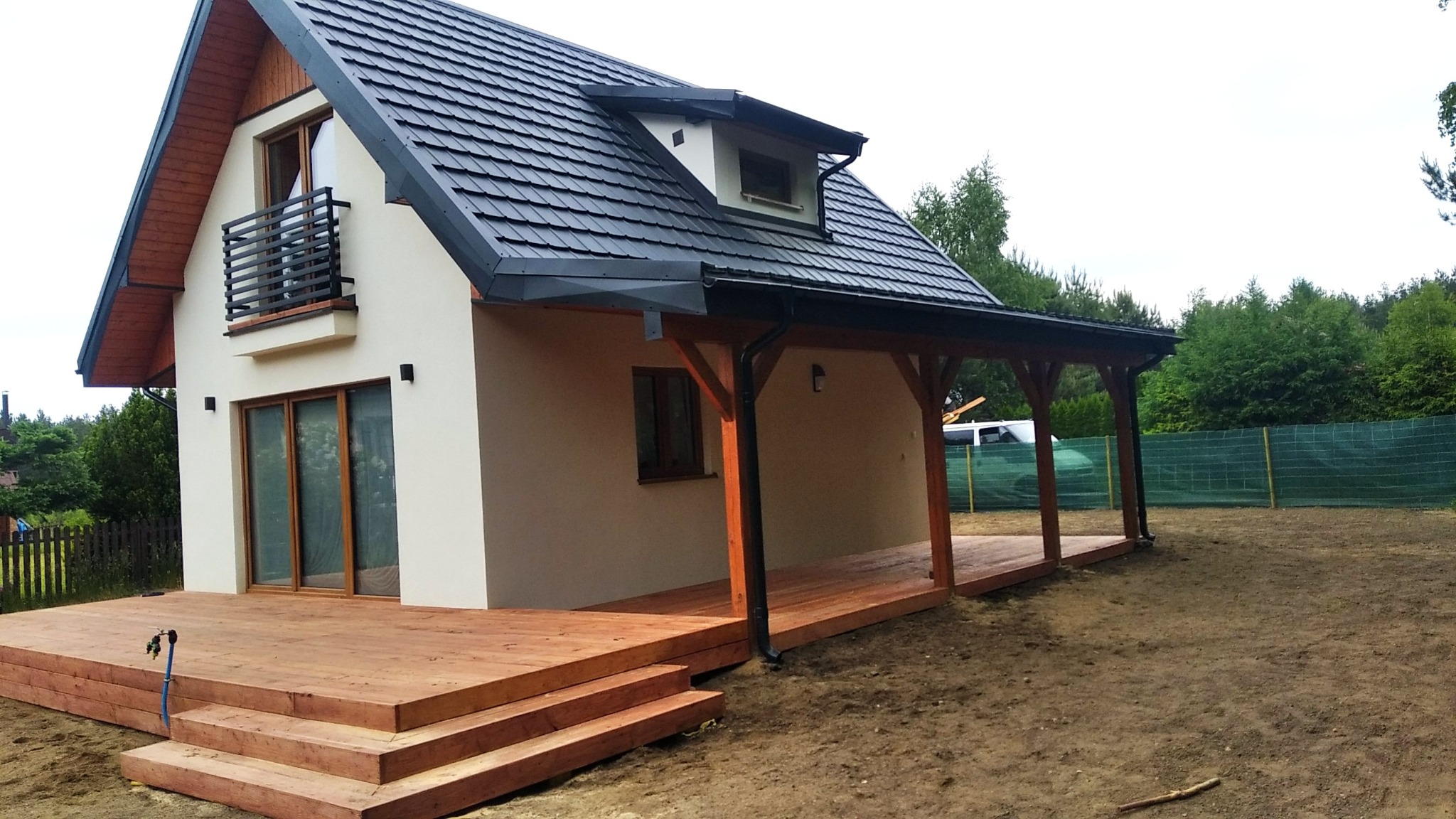 Nowoczesny dom z drewnianym tarasem i zadaszeniem. Elewacja w jasnym odcieniu, ciemna dachówka. Widoczne okna i balkon z metalową balustradą. Otoczenie: trawnik i drzewa.