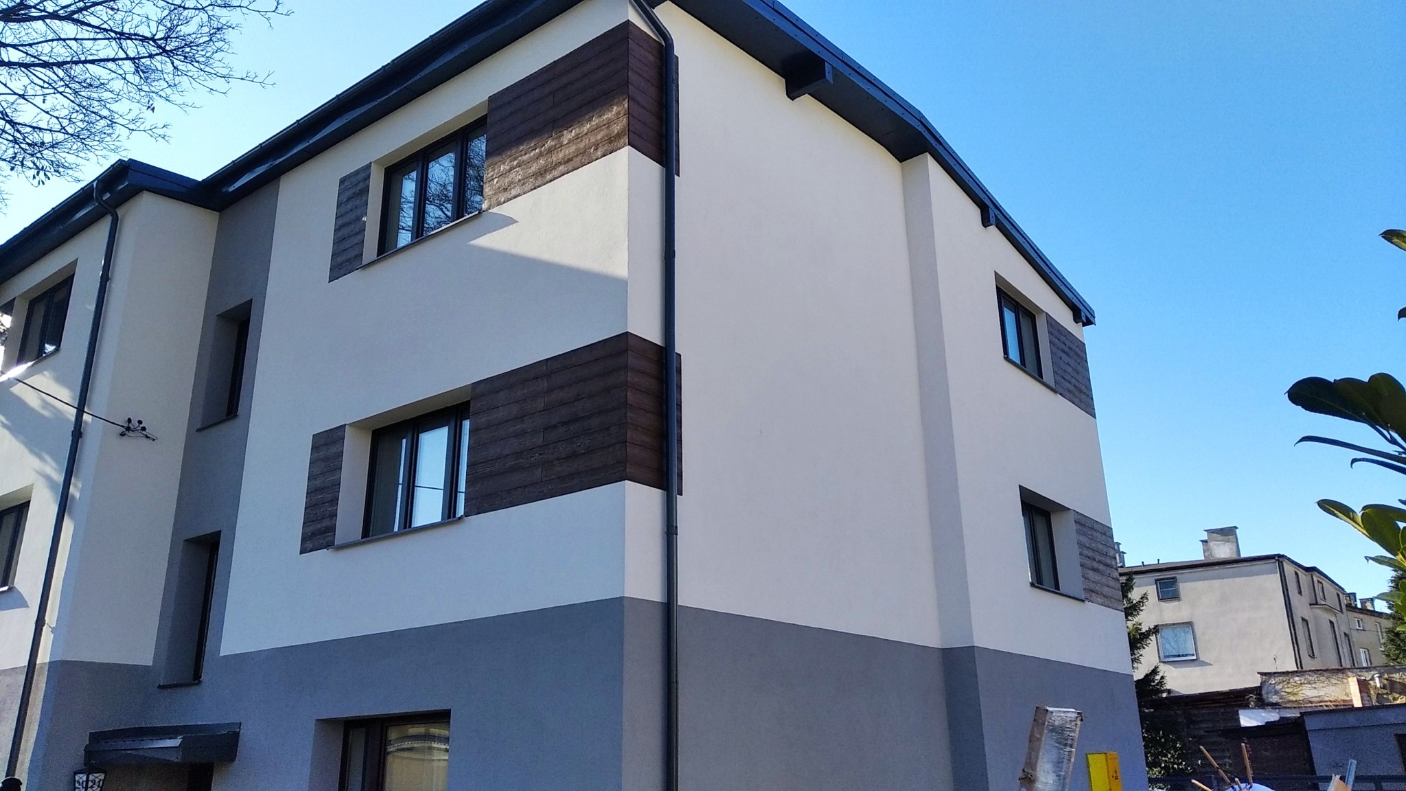 Dwukondygnacyjny budynek po renowacji elewacji, z drewnianymi akcentami wokół okien i szarą dolną częścią, na tle błękitnego nieba.