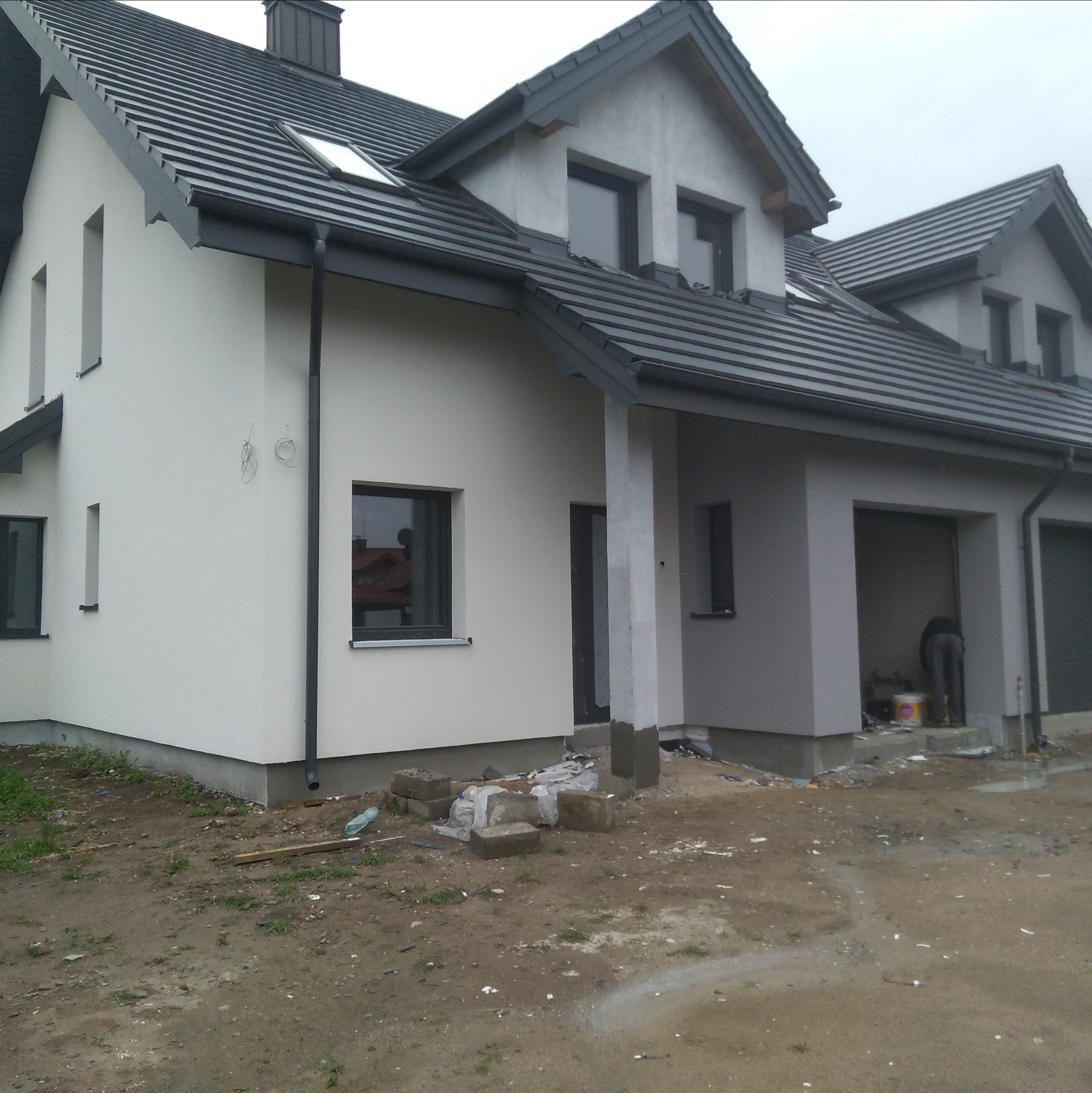 Dwukondygnacyjny dom z szarą dachówką i białą elewacją, częściowo pomalowaną na szaro. Widoczne okna z ciemnymi ramami i teren wokół domu w trakcie prac wykończeniowych.