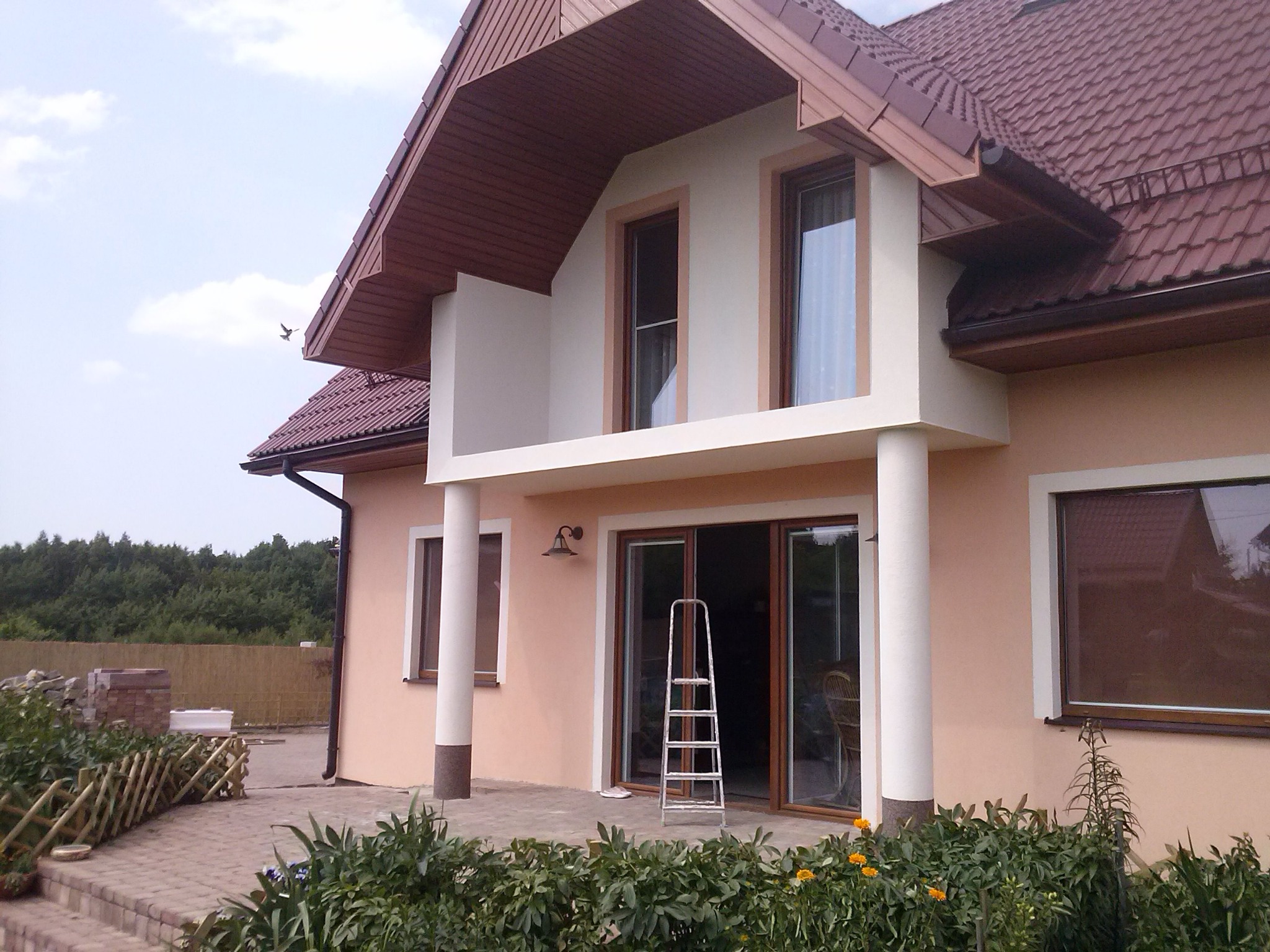 Dom jednorodzinny po renowacji elewacji, z brązowym dachem i beżową fasadą. Na pierwszym planie widoczna drabina oparta o ścianę i fragment ogrodu z niskim płotkiem.