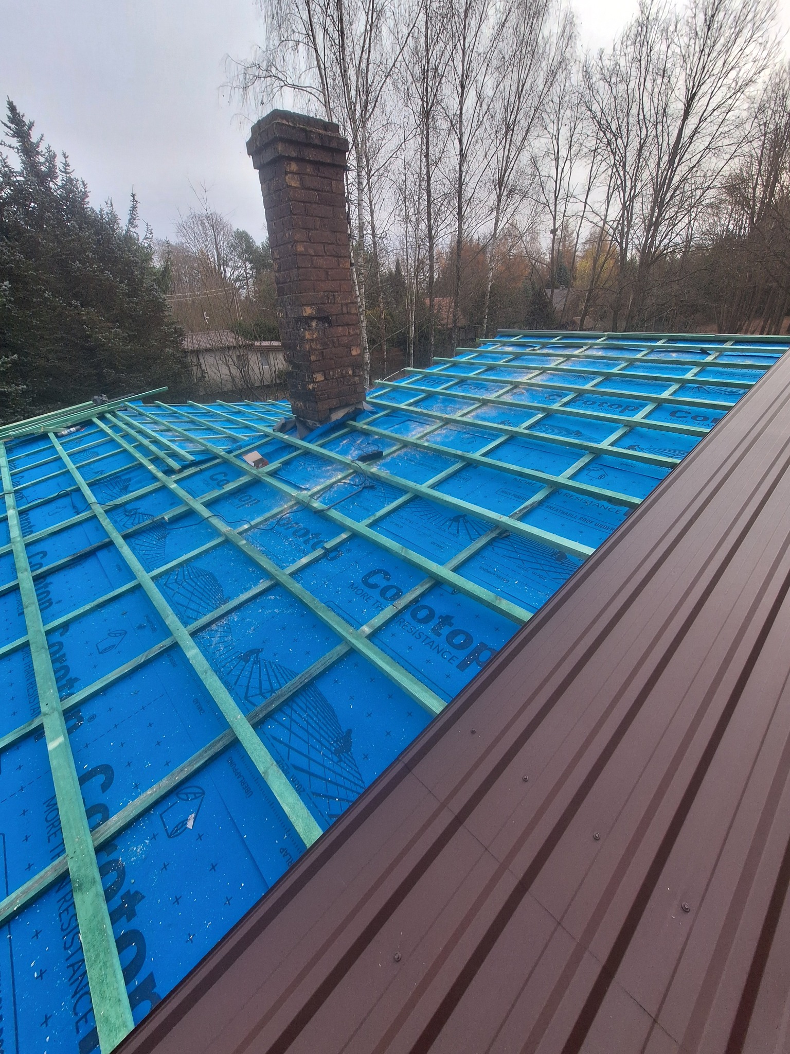 Dach w trakcie budowy: widoczna więźba zaimpregnowana na niebiesko, komin z cegły i fragment brązowego pokrycia. Prace dekarskie w toku, widok z góry.