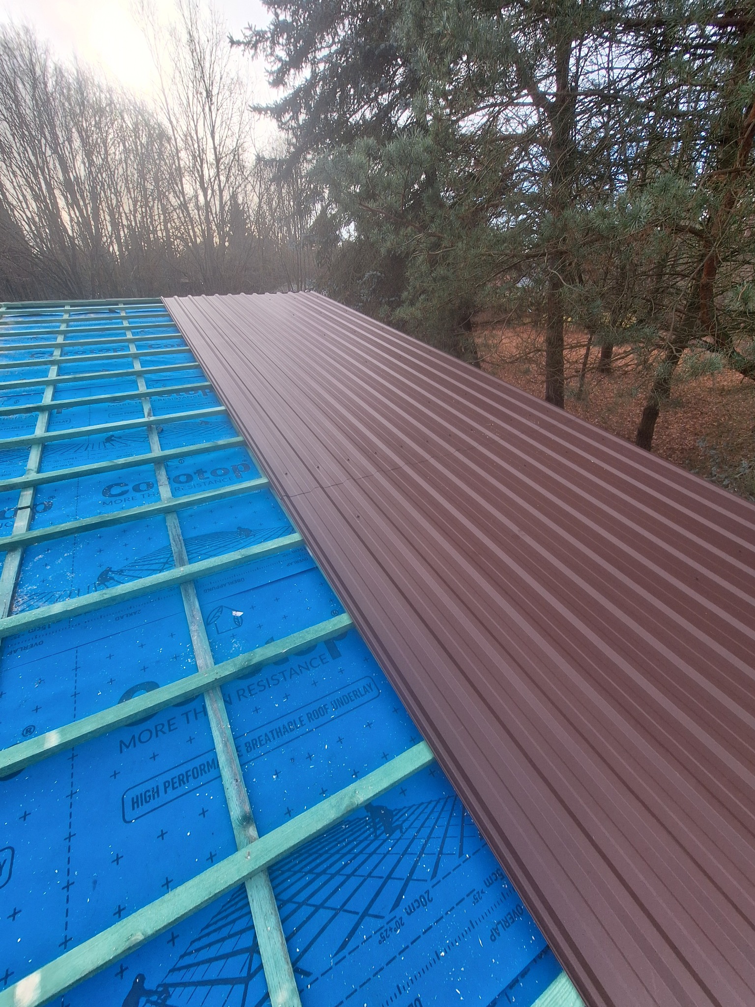 Montaż brązowej blachy trapezowej na dachu z widoczną membraną i drewnianą konstrukcją. Widok z góry, częściowo zalesione tło.