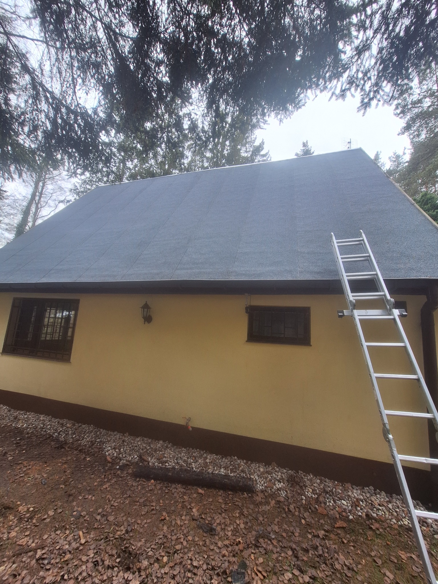 Dom z nowym, ciemnoszarym dachem pokrytym papą. Aluminiowa drabina oparta o ścianę. Widoczne okna z brązowymi ramami i lampa na elewacji.