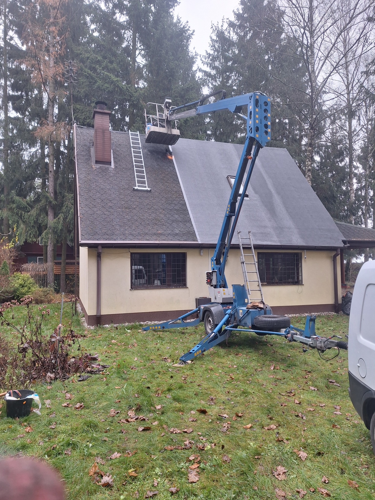 Prace na dachu domu jednorodzinnego z użyciem podnośnika koszowego. Widoczna drabina oparta o komin i fragment elewacji. Prace wysokościowe w jesiennej scenerii.