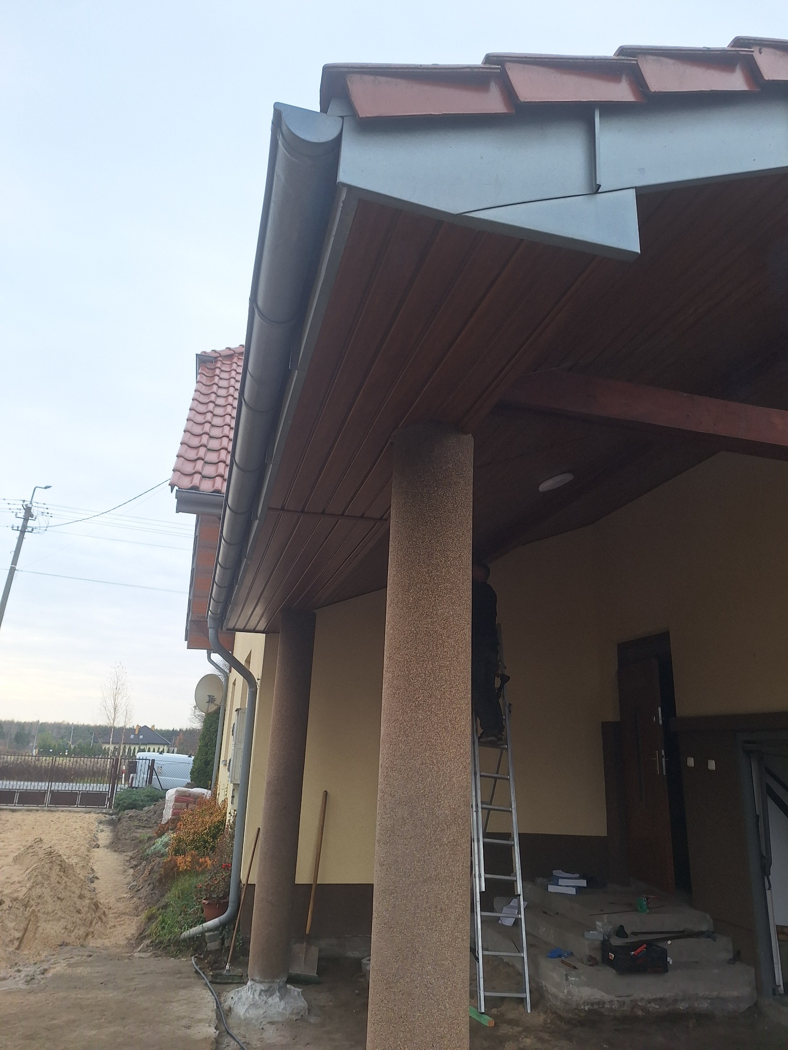 Montaż rynny na brązowym podbitku dachowym domu jednorodzinnego. Widoczna drabina i narzędzia. Ujęcie z dołu, podkreślające detale wykończenia.