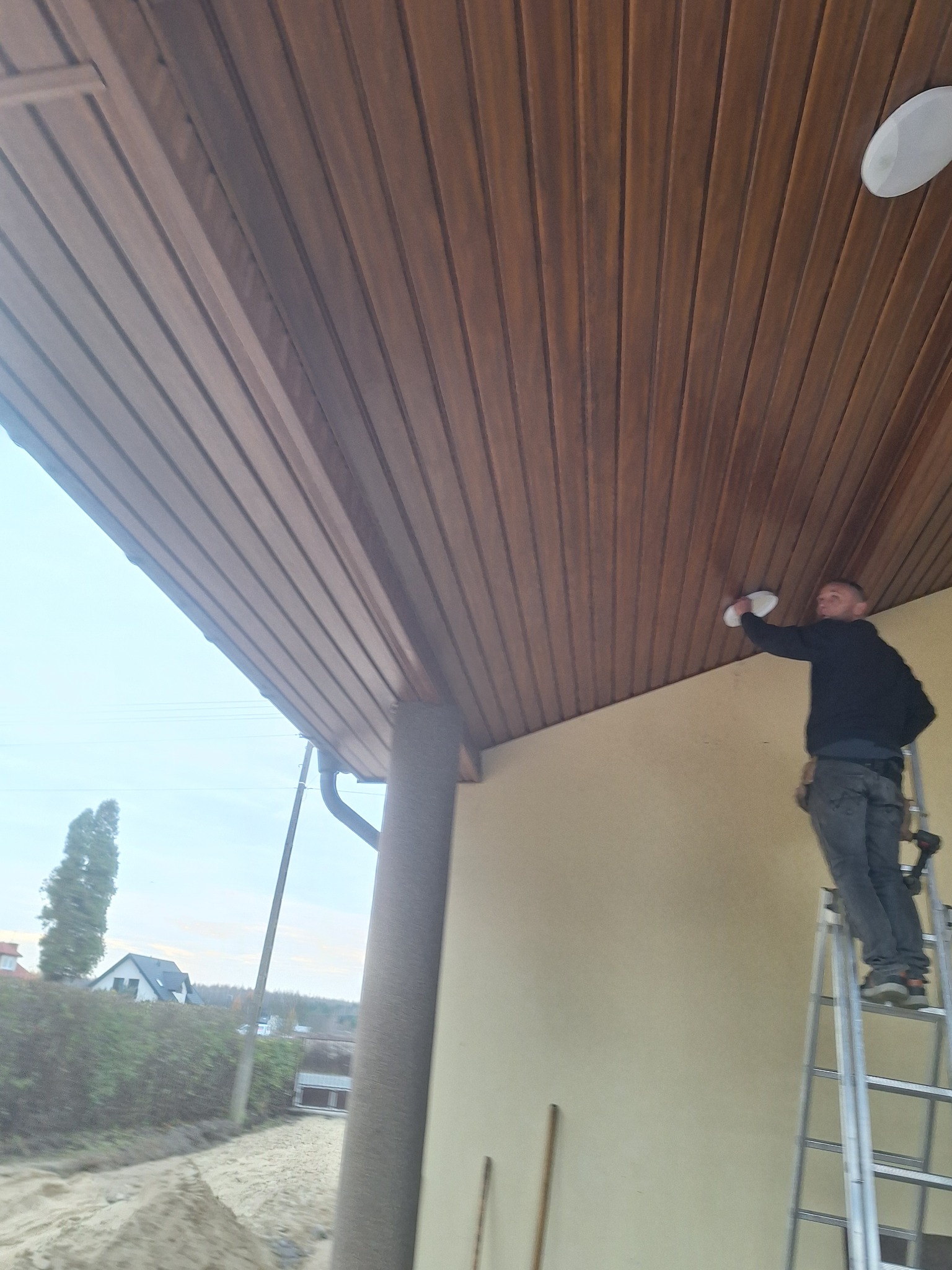 Pracownik na drabinie montuje oświetlenie na drewnianej podbitce dachowej. Widoczne narzędzia i fragment elewacji budynku. Ujęcie z dołu.