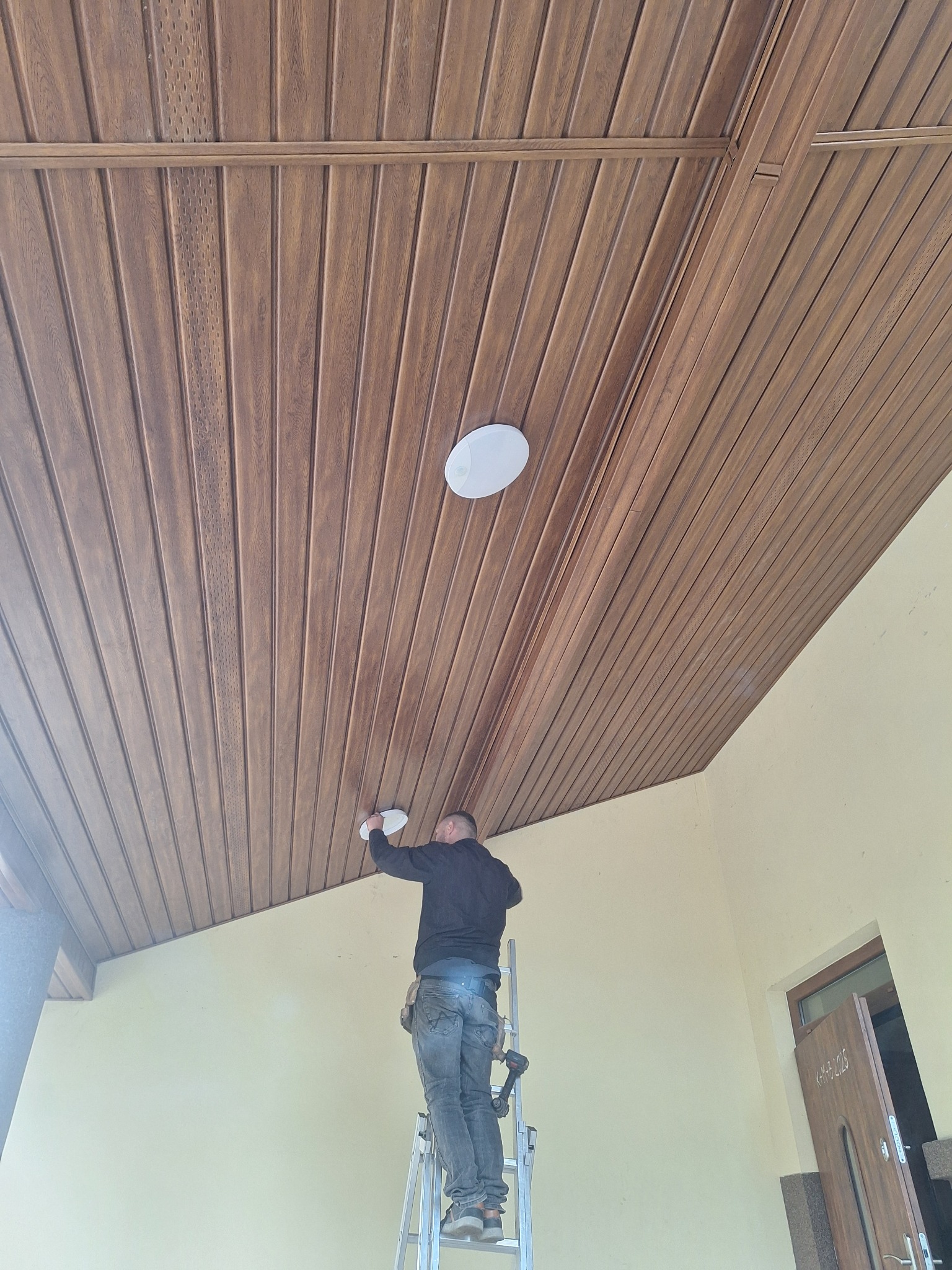 Montaż oświetlenia na drewnianej podbitce dachowej przez pracownika na drabinie. Widok z dołu, jasne wnętrze.