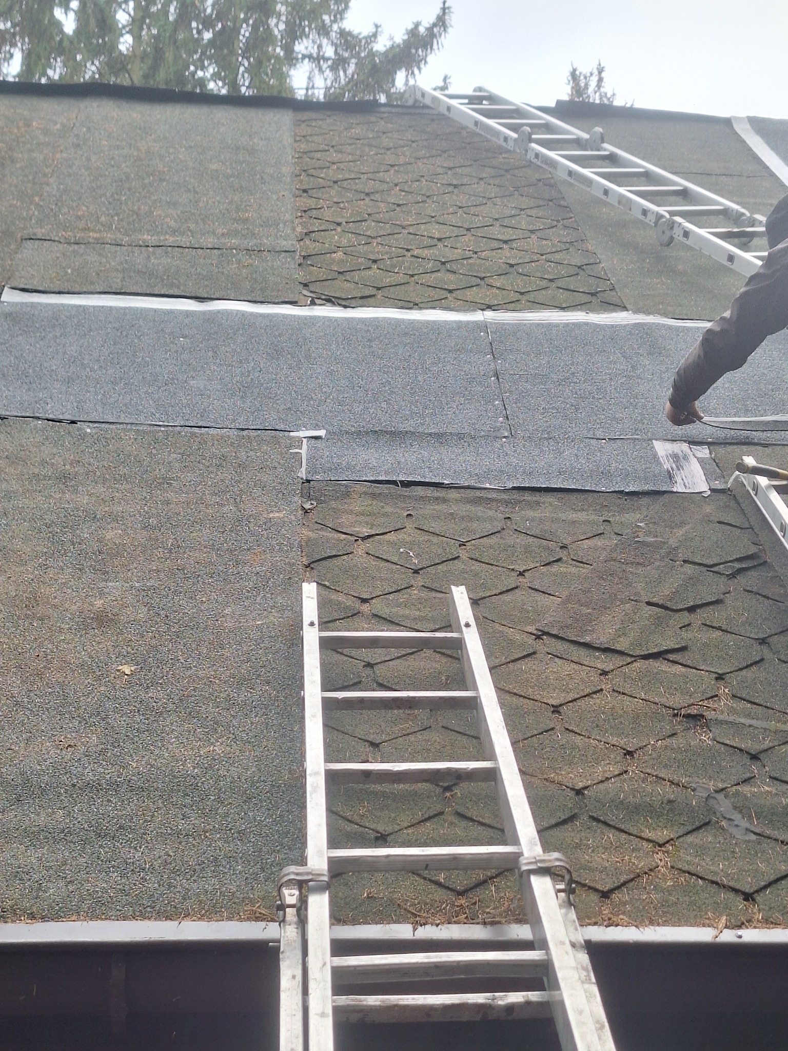 Fragment dachu z bitumiczną papą, częściowo pokryty nową warstwą. Dwie drabiny opierają się o dach. Widoczna ręka pracownika podczas montażu. Ujęcie z dołu.