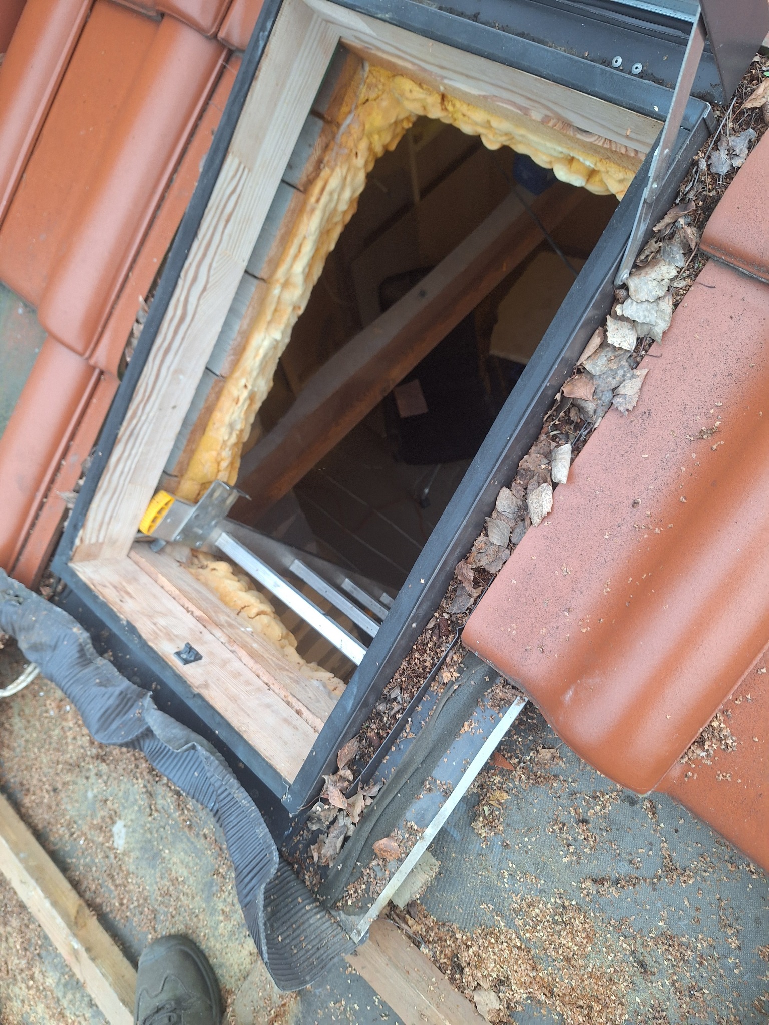 Montaż okna dachowego w dachu z czerwonej dachówki. Widoczne ocieplenie pianką, drewniana rama i elementy mocujące. Na dachu leżą liście i trociny.