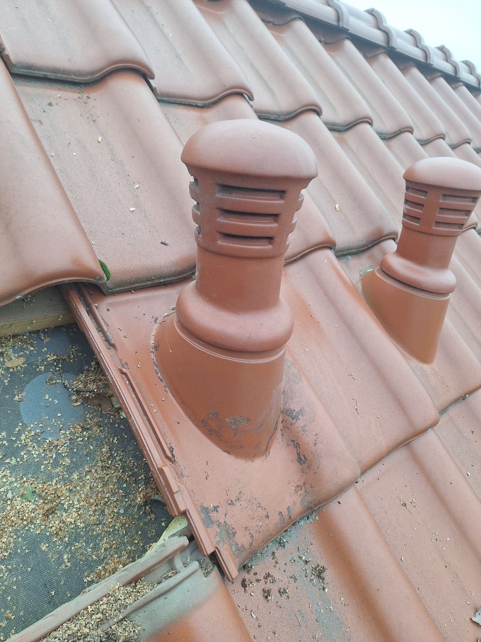 Czerwona dachówka ceramiczna z dwoma kominkami wentylacyjnymi. Widoczne zabrudzenia i liście na powierzchni dachu. Ujęcie z bliska, ukazujące detale.