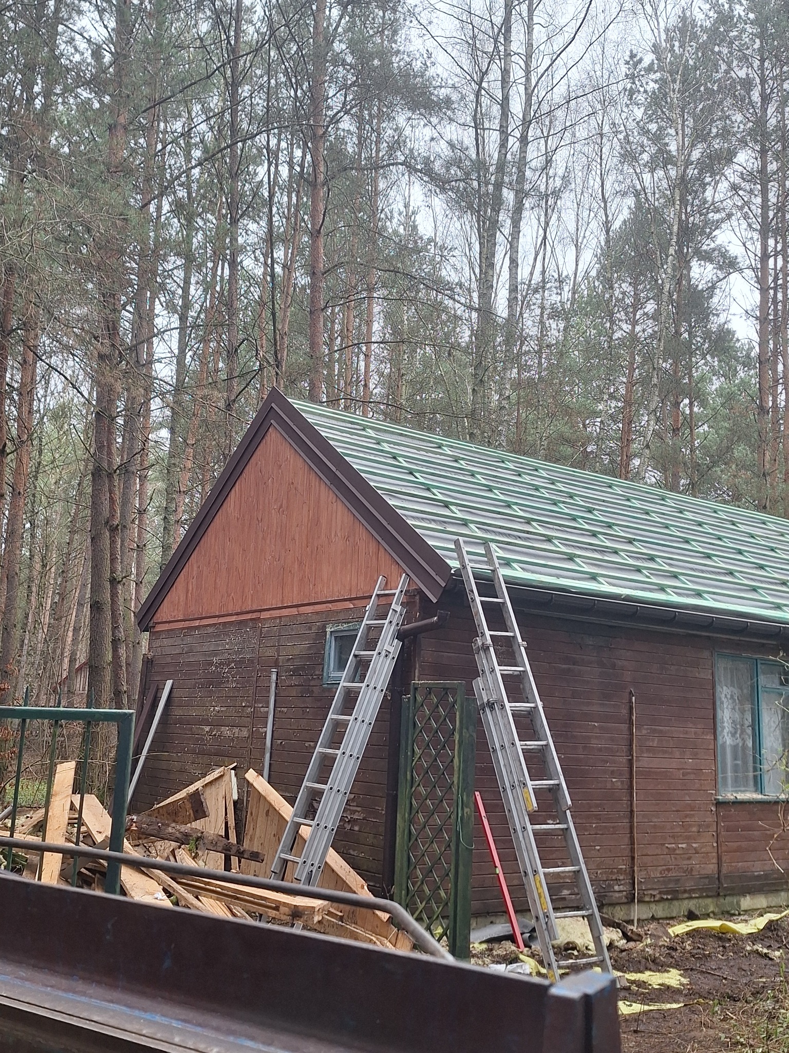 Budynek z nowym, zielonym dachem na tle lasu. Dwie drabiny opierają się o ścianę. Widoczne resztki materiałów budowlanych i fragment rynny. Ujęcie z dołu.