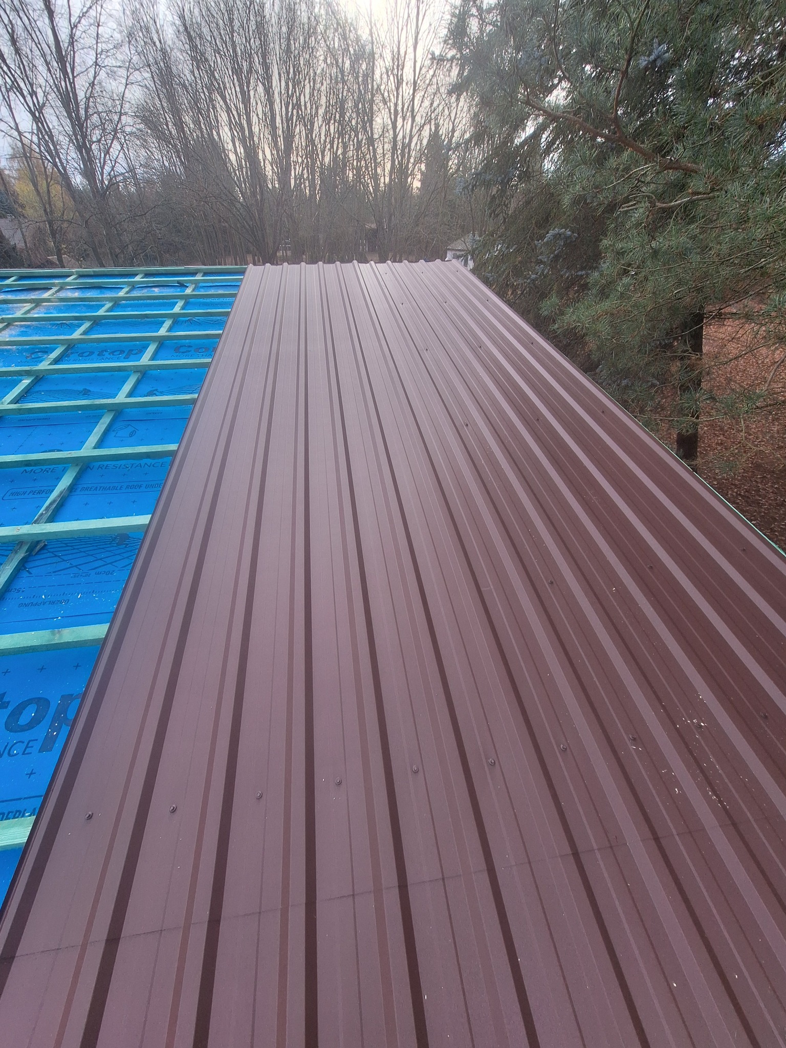 Montaż brązowego dachu z blachy trapezowej, widoczne elementy konstrukcji i membrany dachowej. Prace dekarskie w toku, zieleń drzew w tle.