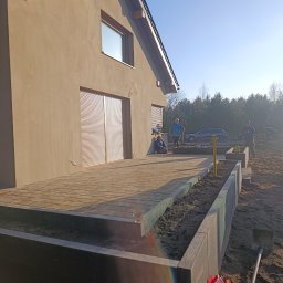 Royal Green Garden Oleksandr Vashchenko - Nowoczesny dom z tarasem z kostki, obrzeża rabat z betonu. W tle osoby i samochód. Widok na ogród w trakcie prac ziemnych, narzędzia ogrodnicze. Słoneczny dzień.