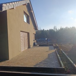Royal Green Garden Oleksandr Vashchenko - Ułożona kostka brukowa na tarasie przy domu z widocznym mini-koparką i pracownikami w tle. Jasne oświetlenie.
