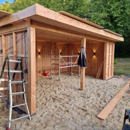 Royal Green Garden Oleksandr Vashchenko - Drewniana konstrukcja altany w trakcie budowy, widoczne rusztowanie, drabina i narzędzia. Na dachu pracuje osoba. Oświetlenie zamontowane. Podłoże piaszczyste.