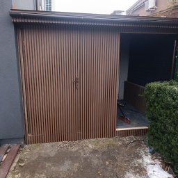 Nowoczesna drewutnia z brązowymi lamelami, zamykana na dwuskrzydłowe drzwi. Wnętrze wyłożone płytami, widoczna miotła. Obok krzew i resztki śniegu.