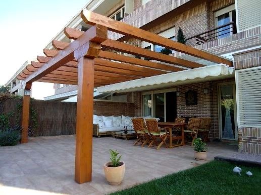 Drewniana pergola nad tarasem z meblami ogrodowymi i roślinami w donicach. Widoczny fragment trawnika i elewacja budynku z cegły.