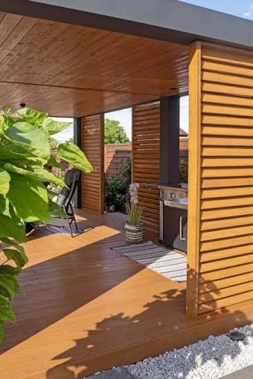 Zadaszony taras z drewnianą podłogą i ścianami z żaluzji, wyposażony w grill. W tle widoczny ogród i drewniane ogrodzenie. Nowoczesny design.