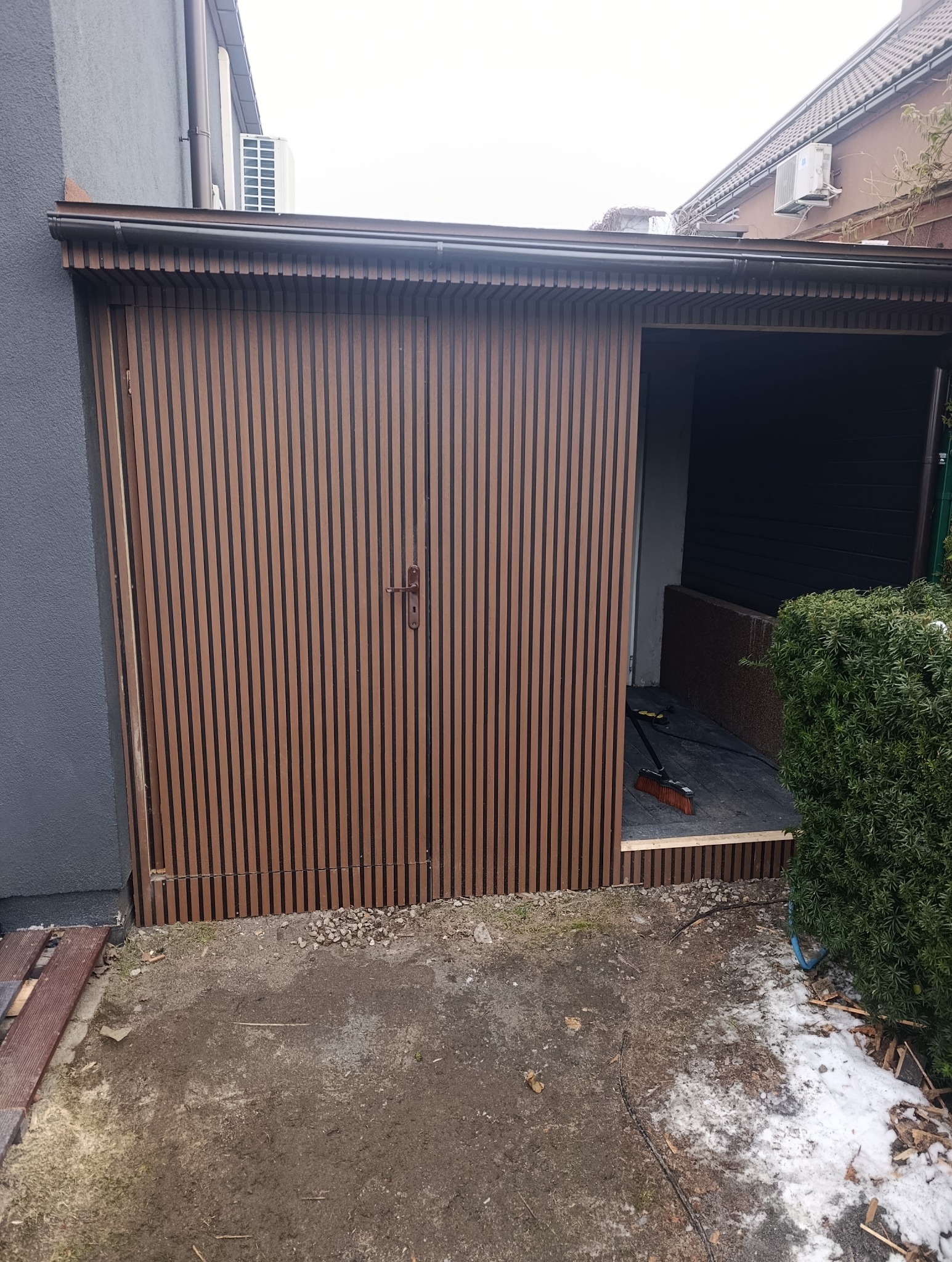 Nowoczesna drewutnia z brązowymi lamelami, zamykana na dwuskrzydłowe drzwi. Wnętrze wyłożone płytami, widoczna miotła. Obok krzew i resztki śniegu.