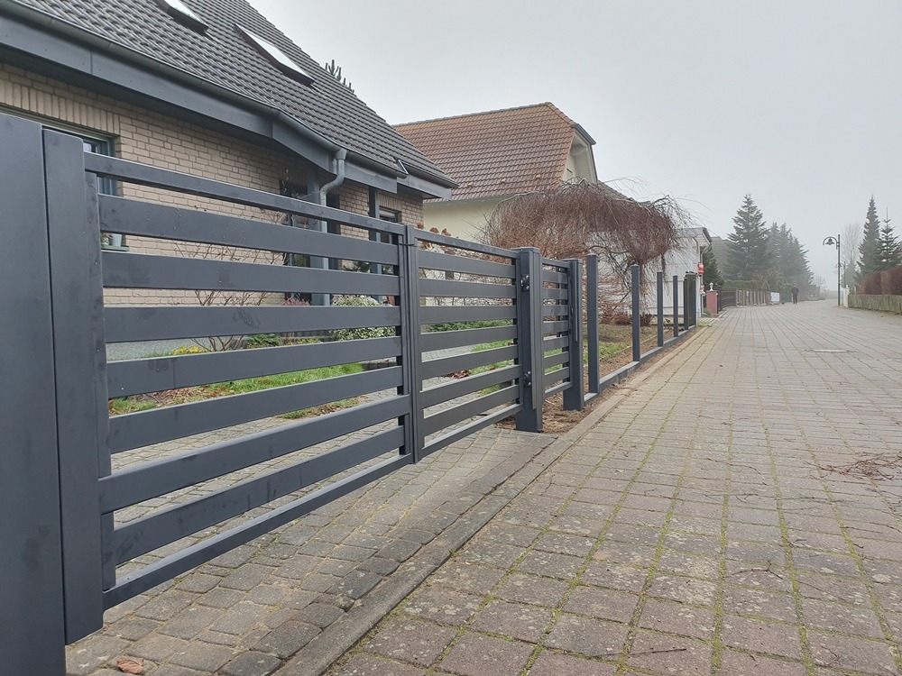 Nowoczesne, poziome ogrodzenie panelowe w kolorze grafitowym, oddzielające posesję od chodnika z kostki. Widok na dom i ulicę w tle, pochmurny dzień.