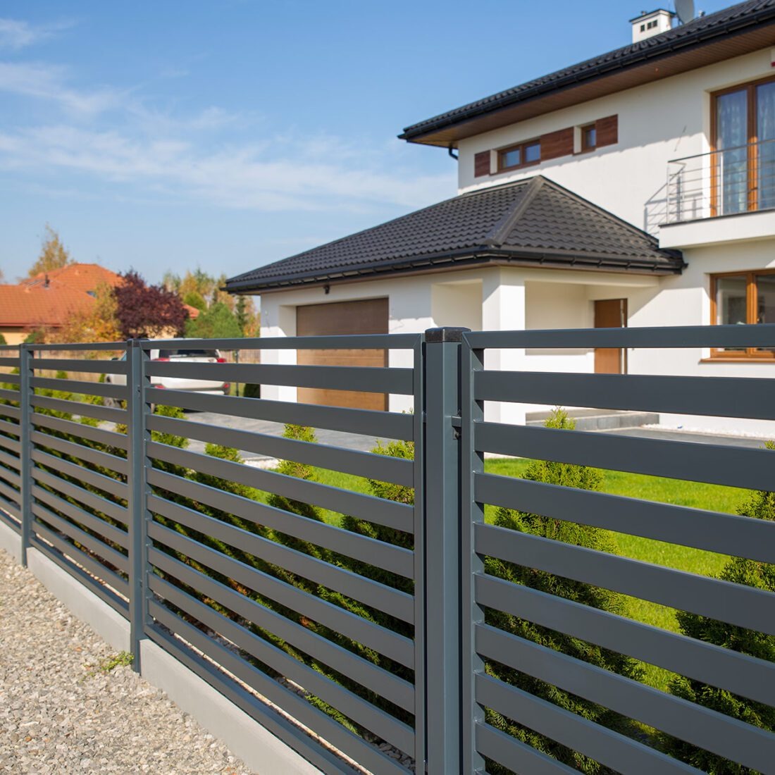 Nowoczesne, poziome ogrodzenie panelowe w kolorze grafitowym, otaczające posesję z białym domem i zielonym trawnikiem w tle. Widok z zewnątrz, w słoneczny dzień.