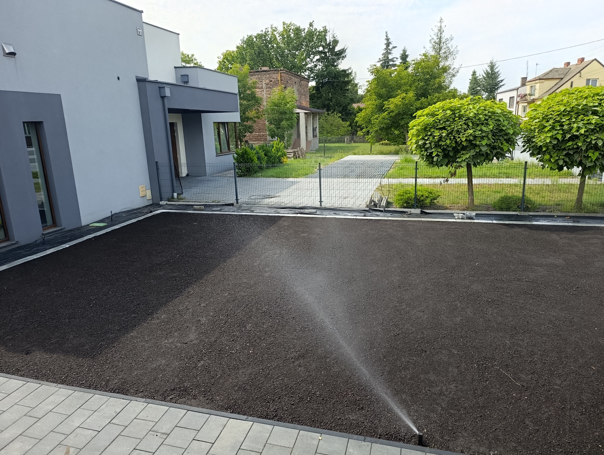 Przygotowany teren pod trawnik z nawadnianiem, widok z góry. Nowoczesny dom z szarą elewacją w tle. Dwa kuliste drzewa ozdobne przy ogrodzeniu.