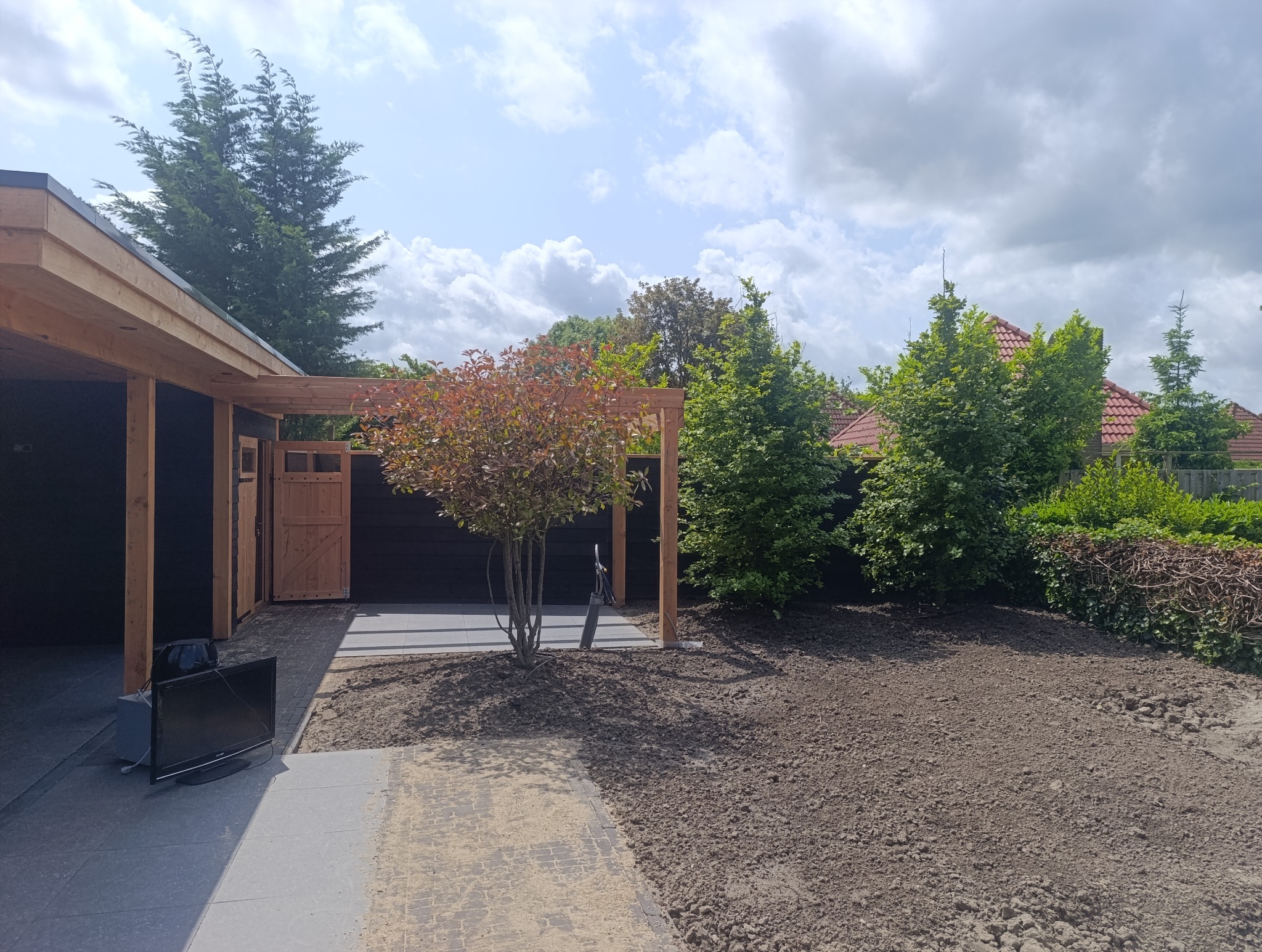 Nowoczesny ogród z drewnianą konstrukcją, świeżo przygotowanym terenem pod trawnik i młodym drzewkiem. W tle widoczne drzewa i zabudowania.