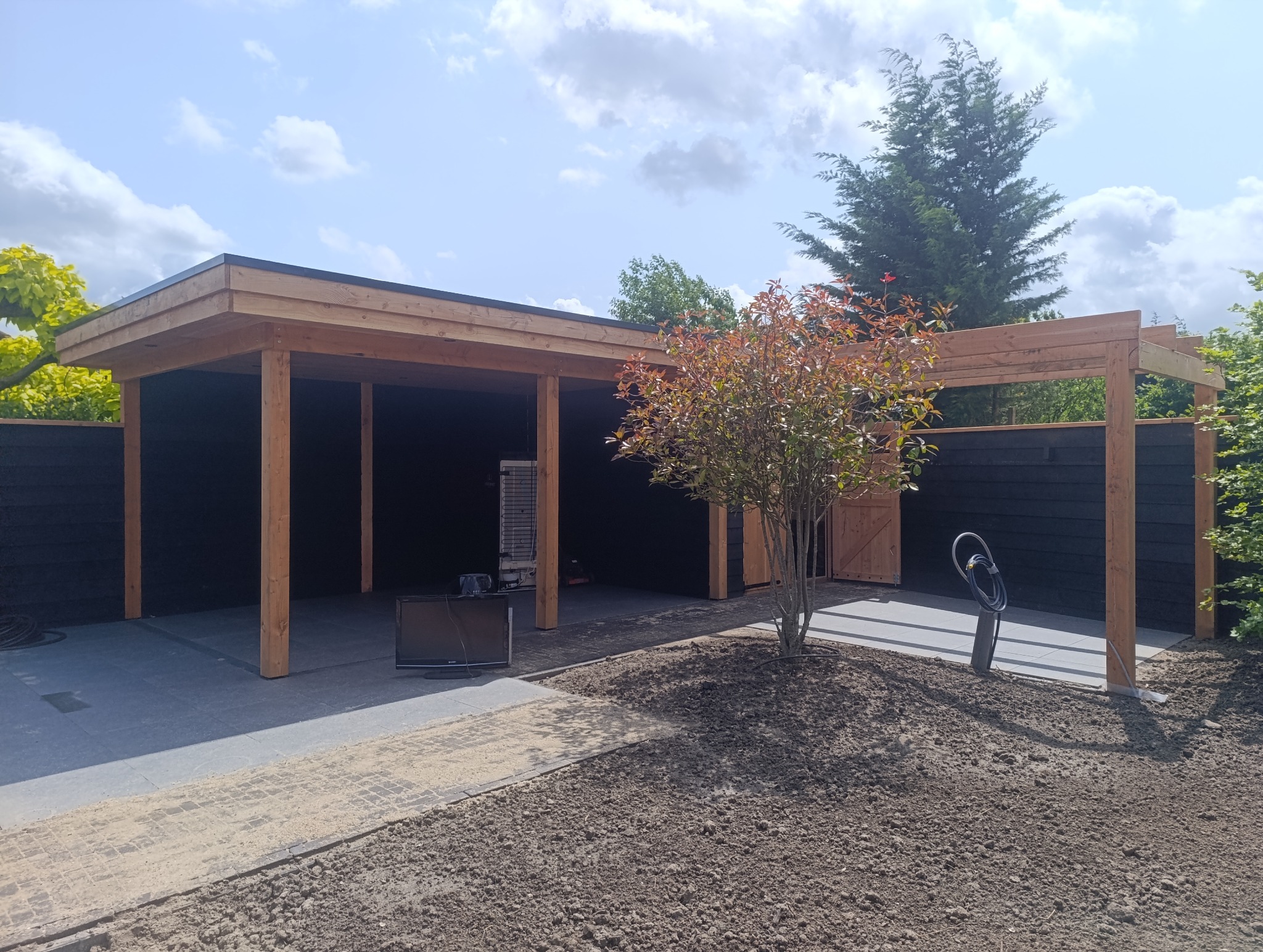 Nowoczesna wiata garażowa z drewna i czarnym ogrodzeniem, otoczona świeżo przygotowanym terenem pod trawnik i młodym drzewkiem. Widoczny wąż ogrodowy i stary telewizor.