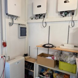 ElektroSquad Krystian Składan - Białe falowniki Huawei zamontowane na ścianie, z widoczną rozdzielnicą i akumulatorem energii. Instalacja w pomieszczeniu gospodarczym z półkami i wyposażeniem.