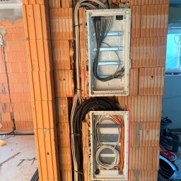 ElektroSquad Krystian Składan - Dwie białe skrzynki rozdzielcze z przewodami elektrycznymi zamontowane w surowej ścianie z cegły dziurawki. Widoczne kable w peszlach i luźne przewody. Etap prac instalacyjnych.