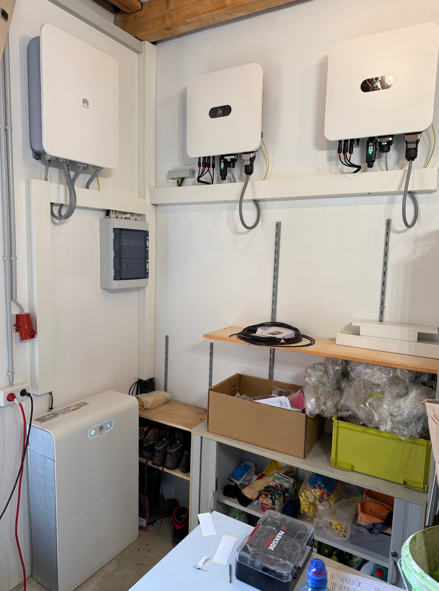 Białe falowniki Huawei zamontowane na ścianie, z widoczną rozdzielnicą i akumulatorem energii. Instalacja w pomieszczeniu gospodarczym z półkami i wyposażeniem.