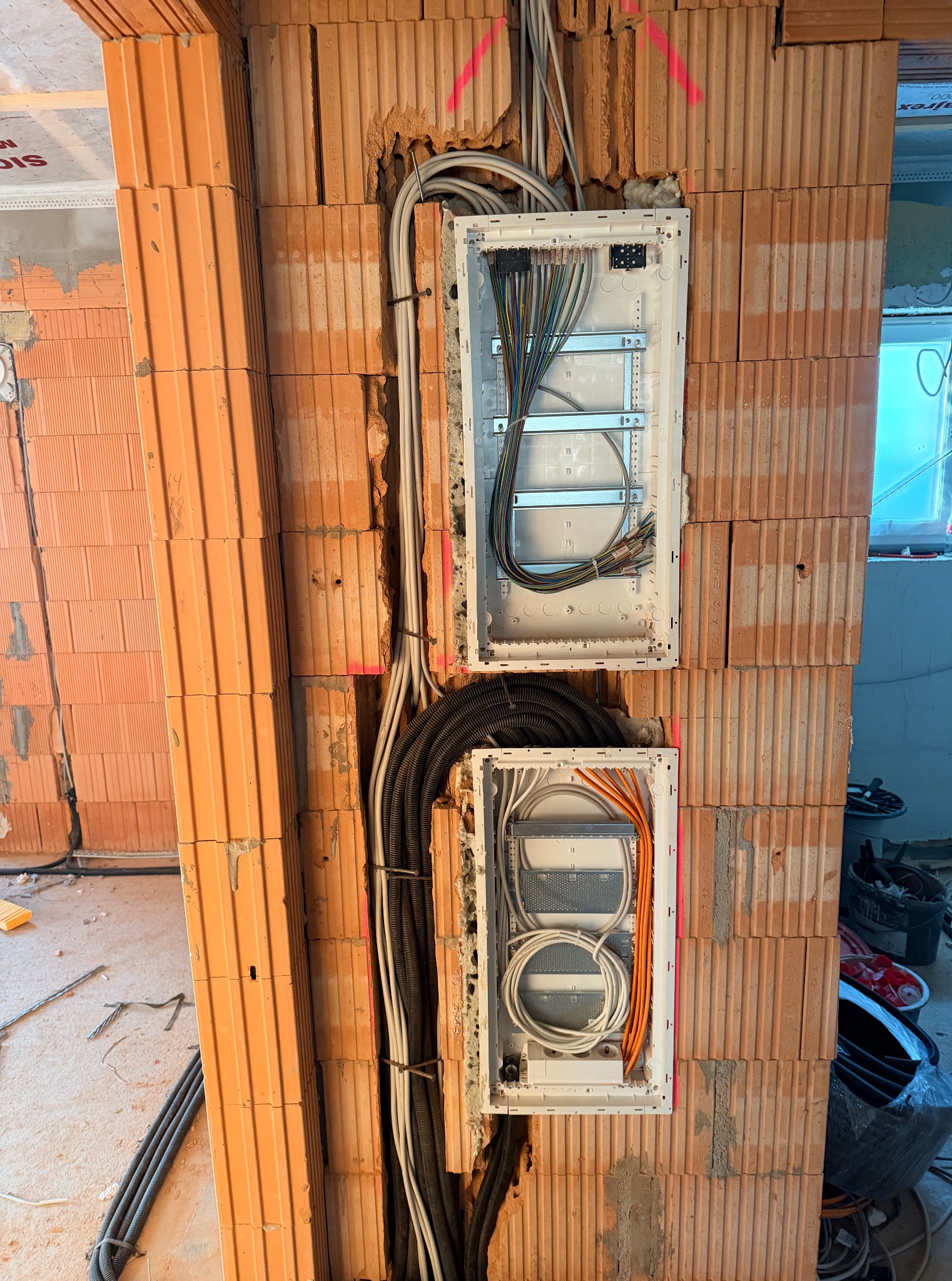 Dwie białe skrzynki rozdzielcze z przewodami elektrycznymi zamontowane w surowej ścianie z cegły dziurawki. Widoczne kable w peszlach i luźne przewody. Etap prac instalacyjnych.
