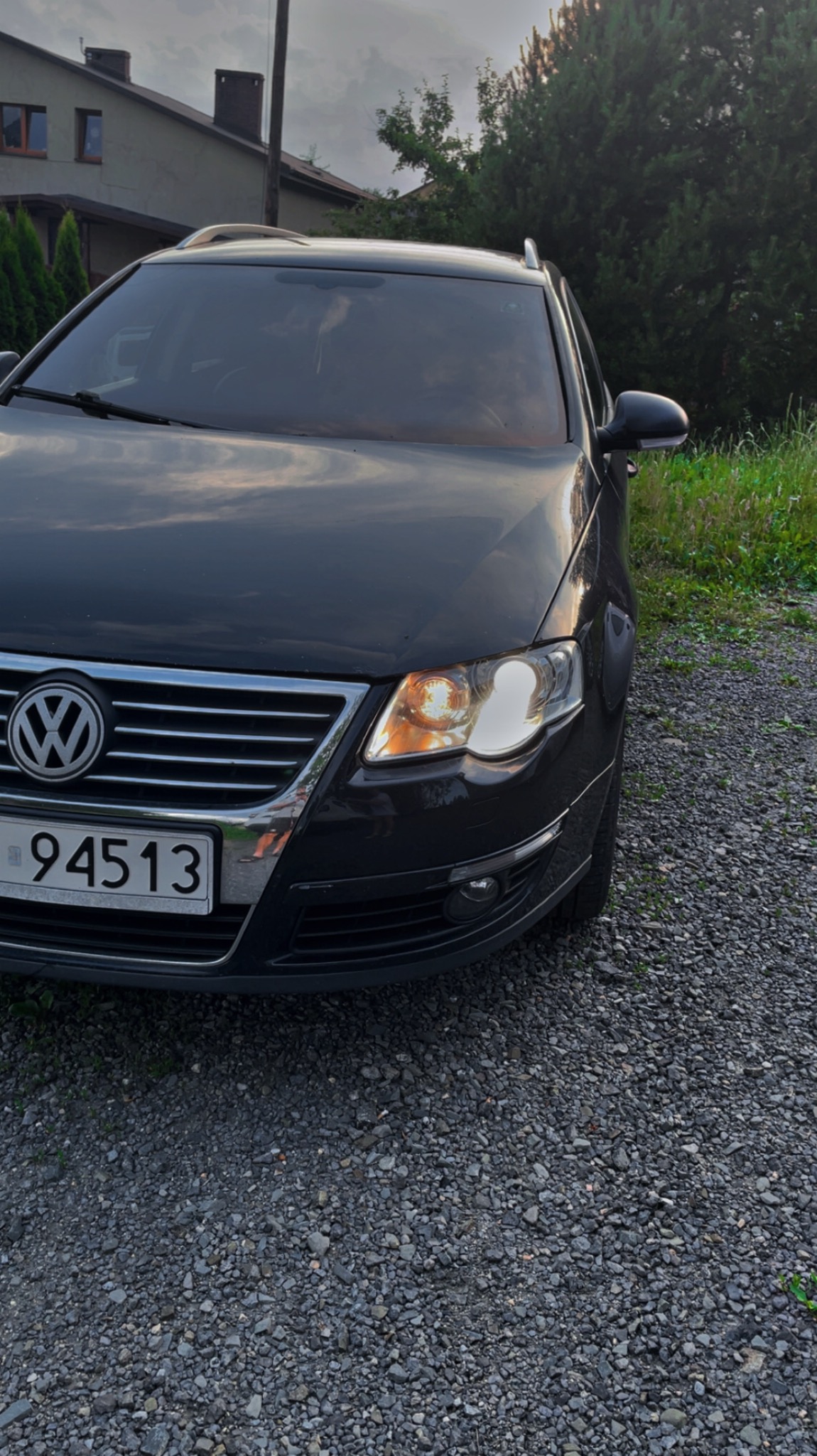 Czarny samochód osobowy Volkswagen Passat z włączonymi światłami mijania, zaparkowany na żwirowym podłożu. Widok z przodu, lekko z boku. W tle budynek mieszkalny i zieleń.