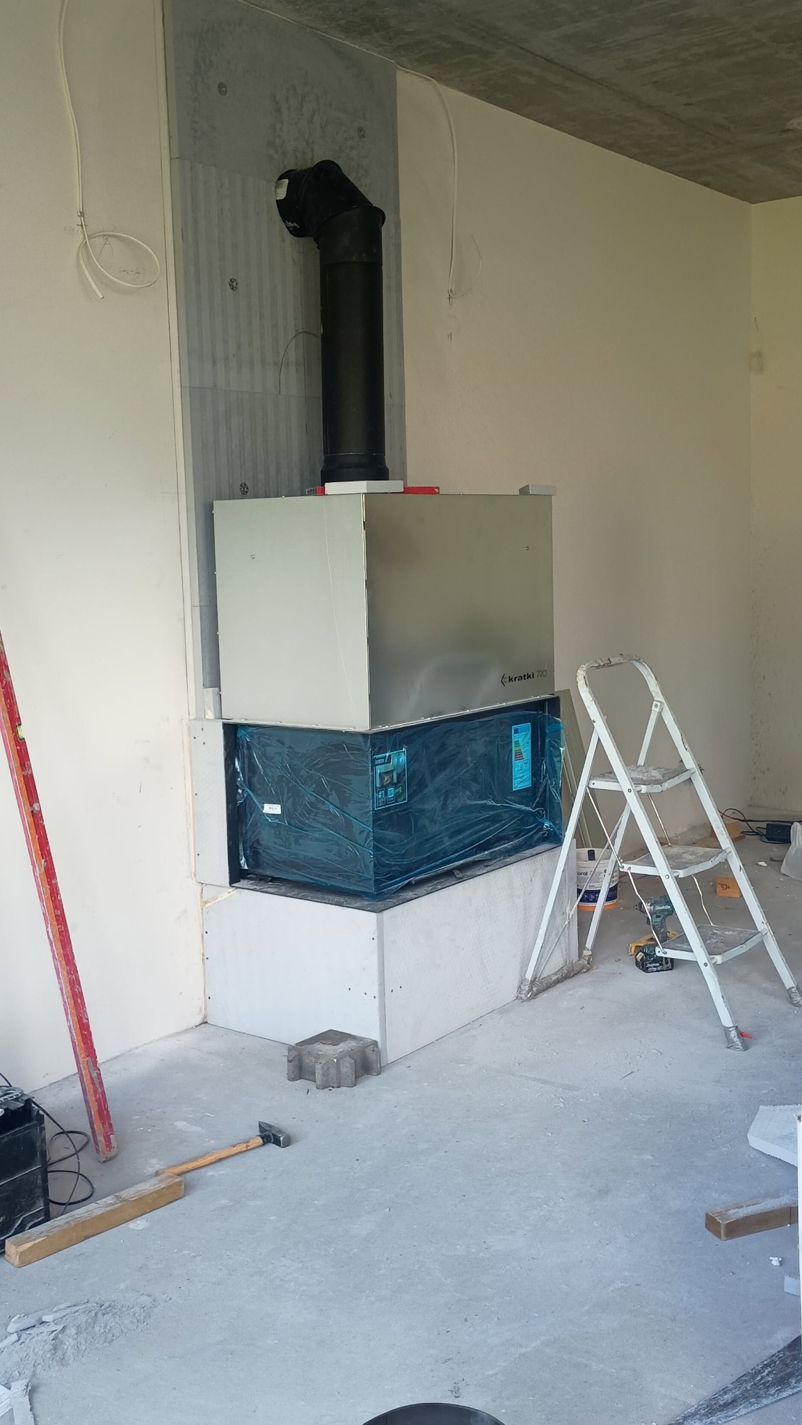 Wnętrze w trakcie budowy kominka z wkładem Kratki, widoczna drabina, poziomica i narzędzia. Wkład kominkowy częściowo zabezpieczony folią. Surowy beton na suficie.