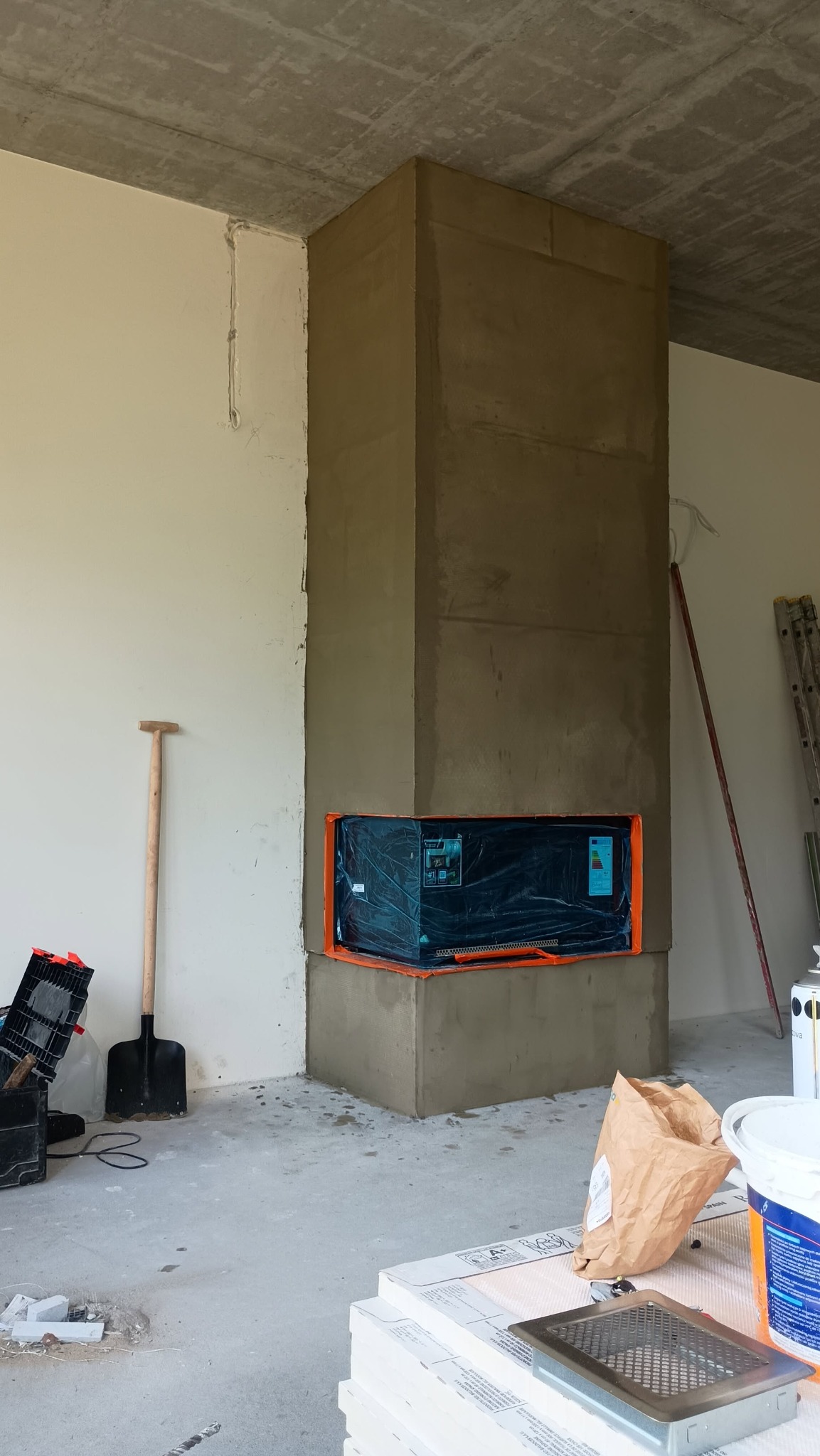 Wnętrze w trakcie prac wykończeniowych: otynkowany komin z wkładem zabezpieczonym folią, narzędzia budowlane, worki z zaprawą i płytki na podłodze.