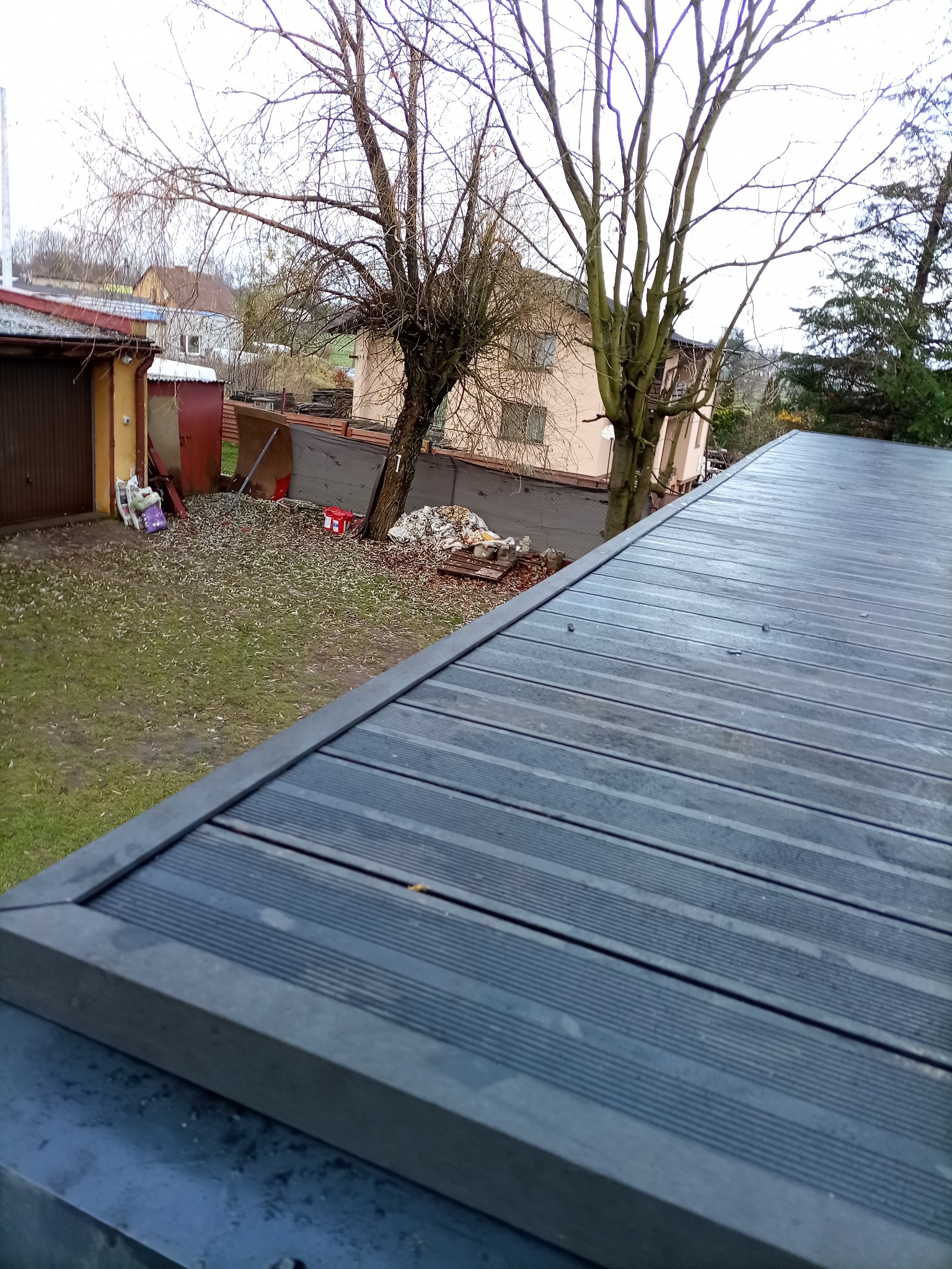 Szary taras kompozytowy z widokiem na podwórze z drzewami i budynkami w tle, uchwycony z góry pod kątem. Deski ułożone poziomo, widoczne mocowania.