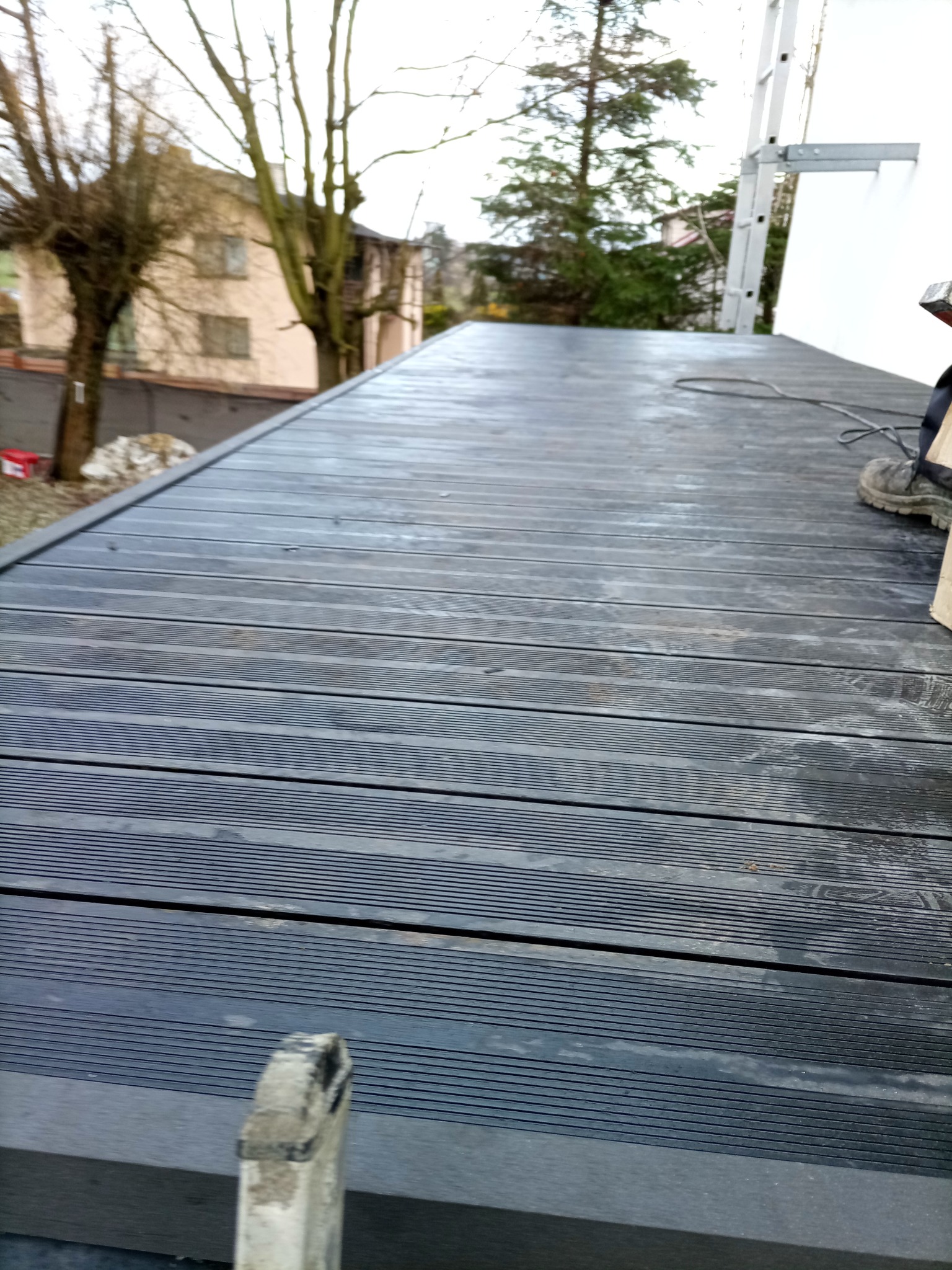 Ciemnoszary taras kompozytowy z widoczną fakturą desek, fragment drabiny w tle, częściowo widoczne buty montera. Ujęcie z niskiej perspektywy.