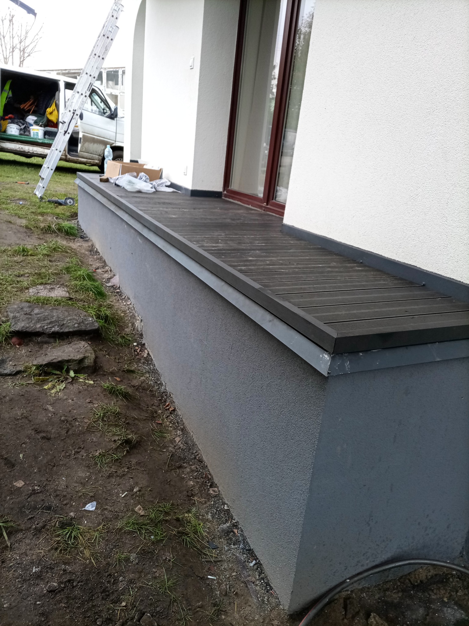 Wykończony taras kompozytowy przy oknie budynku z białą elewacją. Widoczne szare wykończenie ściany i elementy montażowe. W tle drabina i samochód dostawczy.