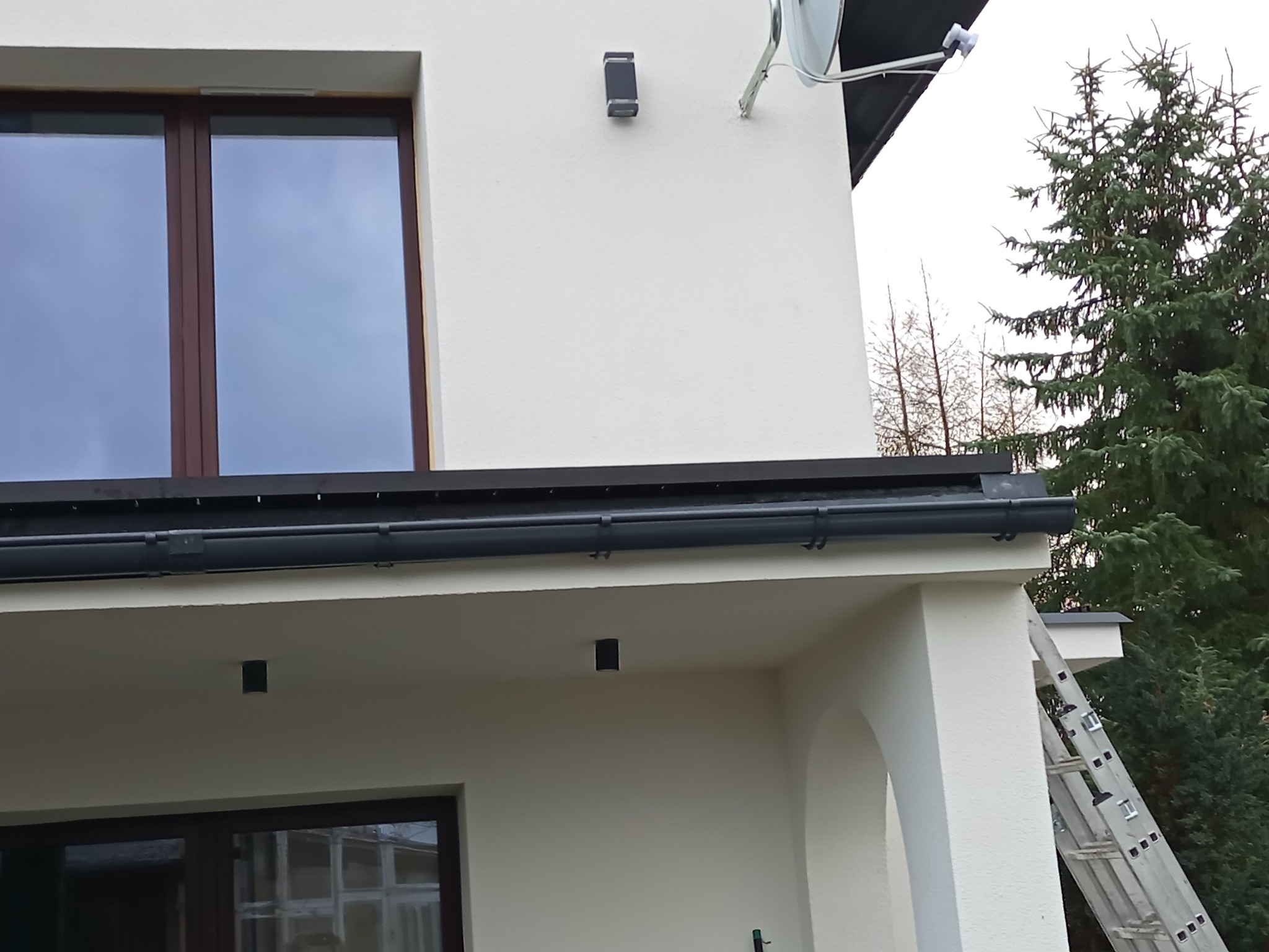 Fragment elewacji domu z oknem, rynnami i lampą zewnętrzną. Widoczna drabina oparta o ścianę. Ujęcie z dołu, podkreślające detale wykończenia.