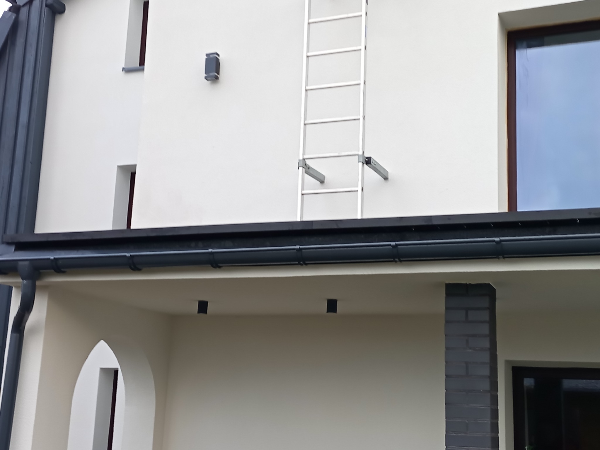 Fragment elewacji budynku z zamontowaną drabinką fasadową, widoczne rynny i elementy oświetlenia zewnętrznego. Ujęcie z dołu, podkreślające detale wykończenia.