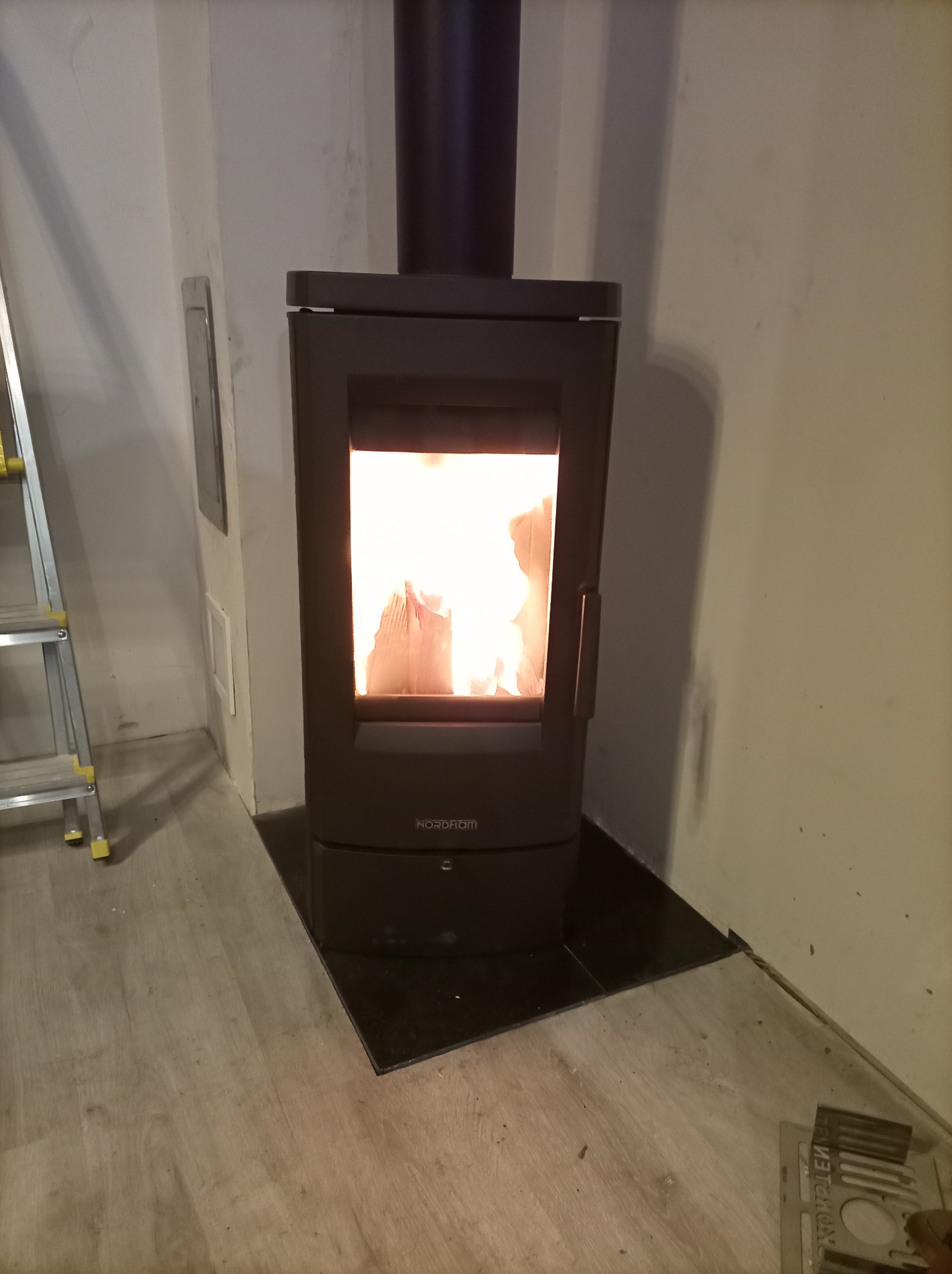 Czarny piec wolnostojący Nordflam z palącym się ogniem, ustawiony na czarnych płytkach, widoczny fragment drabiny i podłogi z paneli.