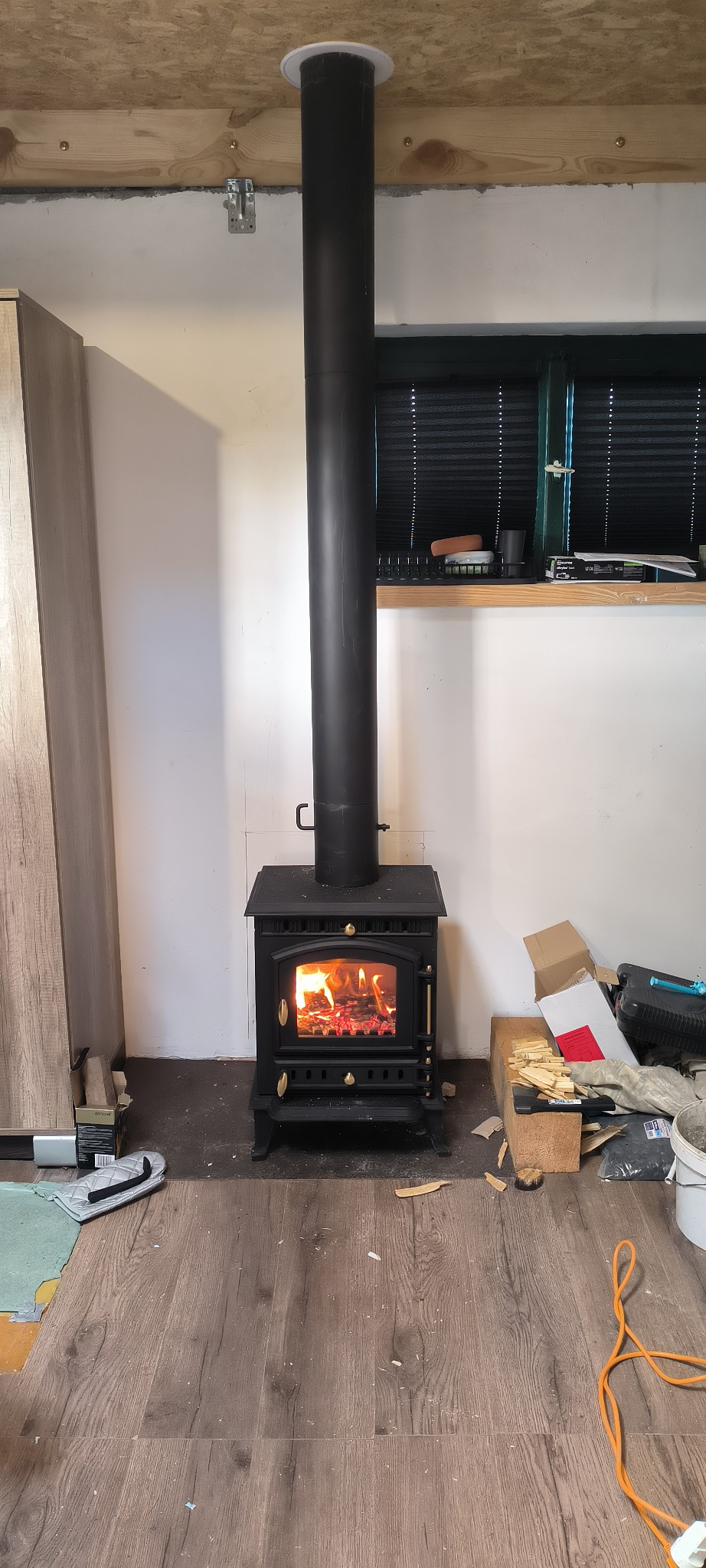 Czarny piec wolnostojący z rurą odprowadzającą dym, zamontowany w pomieszczeniu z panelami podłogowymi i oknem z żaluzjami. Widoczne akcesoria i drewno do palenia.