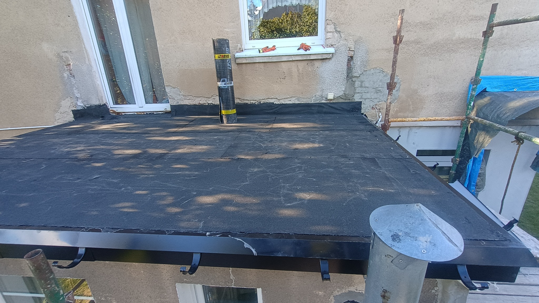 Wykończony płaski dach pokryty czarną papą, z widoczną rolką papy na powierzchni. Rynna w kolorze czarnym. Widoczne okno i fragment elewacji budynku.