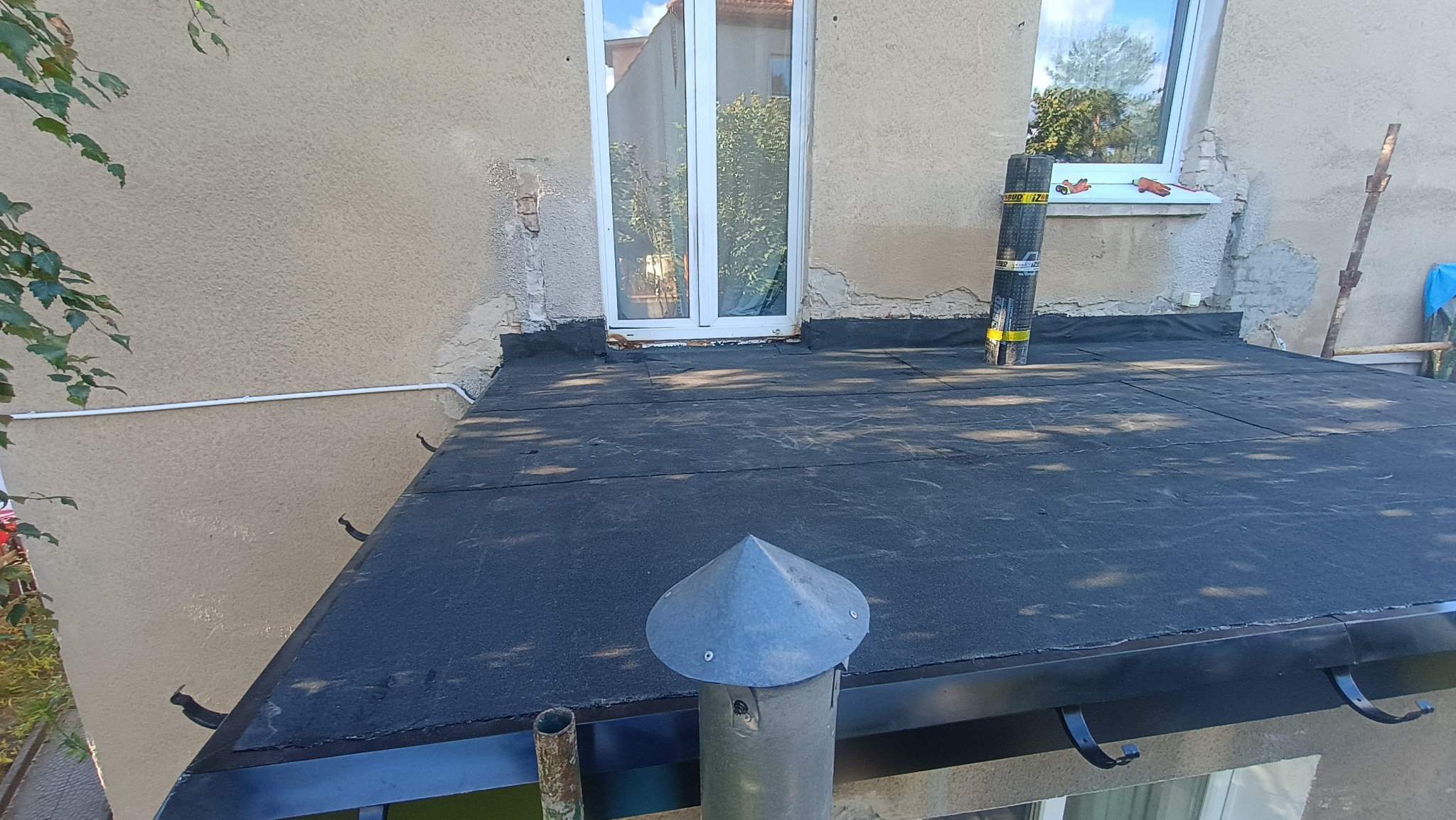 Czarny, płaski dach pokryty papą, z kominem wentylacyjnym i oknem w tle. Widoczna rolka papy na dachu. Detal obróbki blacharskiej przy ścianie budynku.