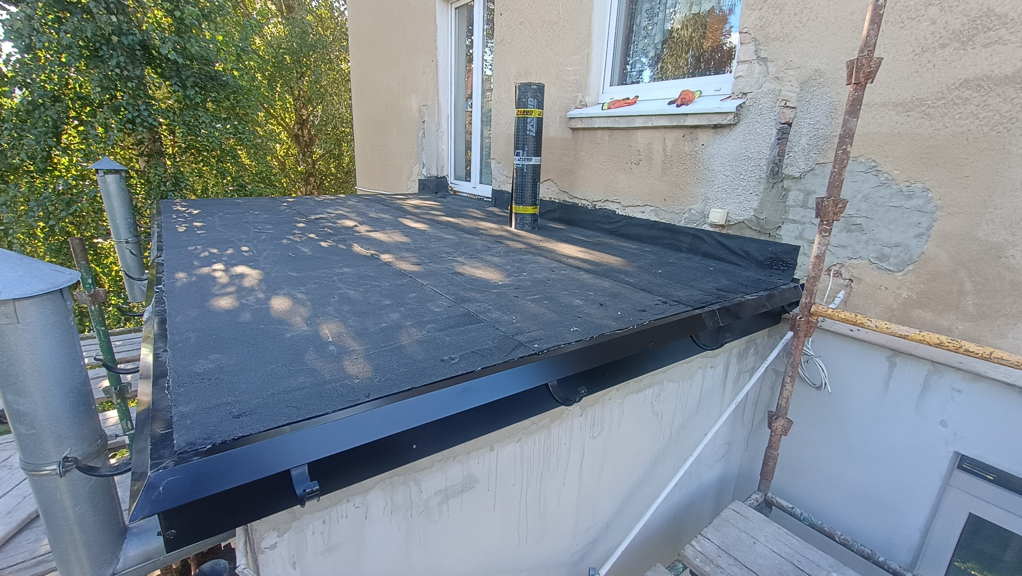 Płaski dach pokryty czarną papą, z zamontowaną czarną rynną. Na dachu stoi rolka papy. W tle fragment elewacji budynku i rusztowanie.