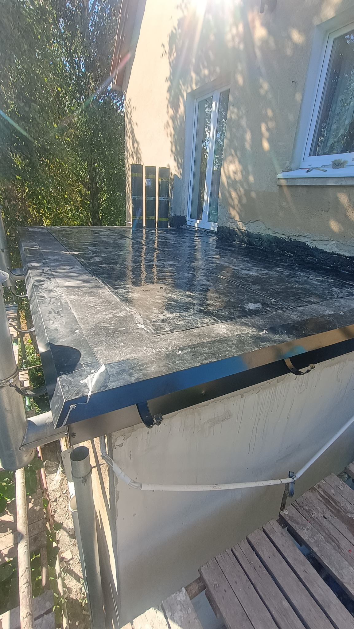 Wykończony płaski dach z czarną papą i obróbkami blacharskimi, widok z rusztowania. Na dachu rolki papy. Słoneczny dzień.