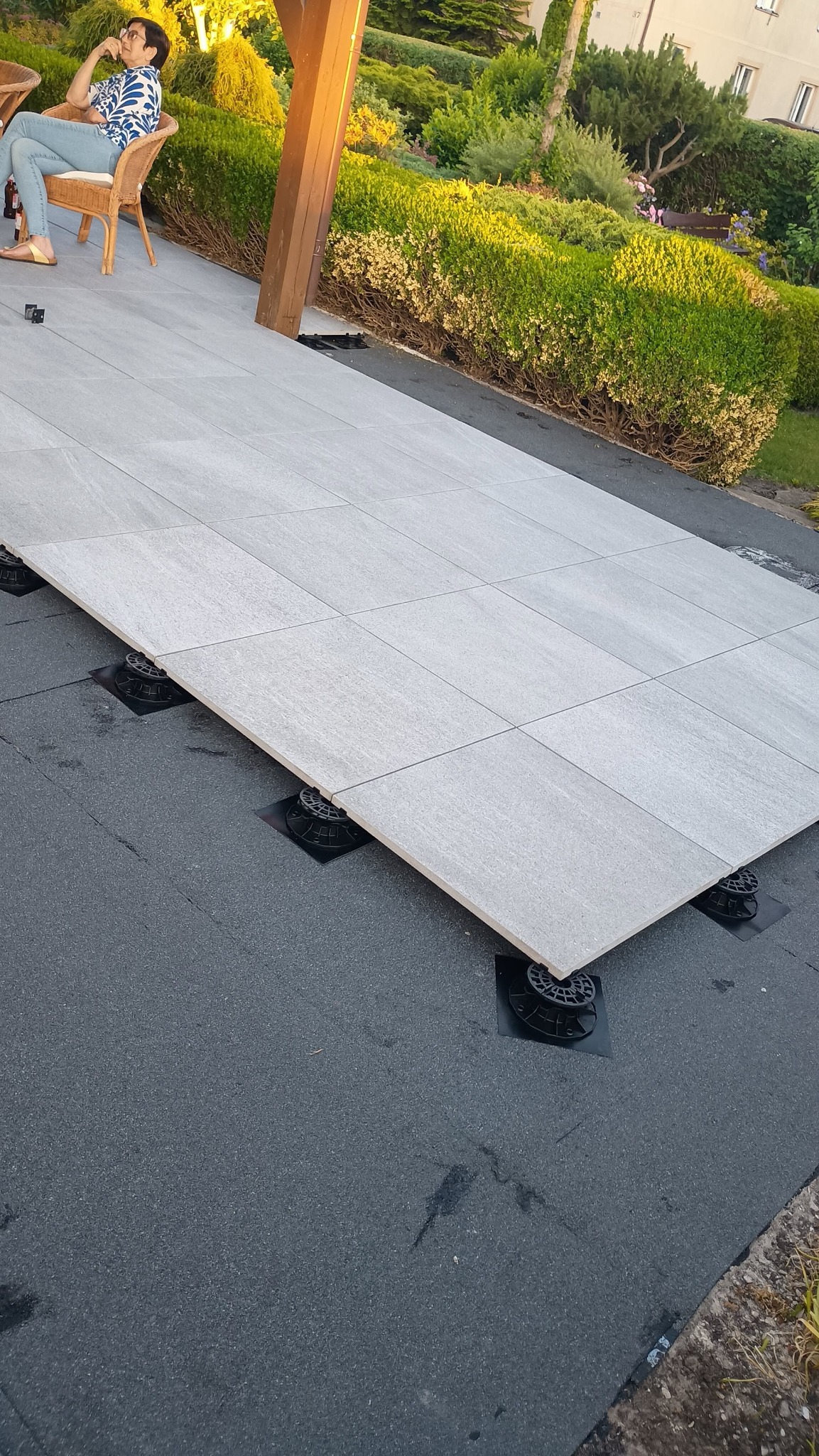 Jasnoszary taras z płyt na regulowanych wspornikach, widok z góry. W tle siedząca osoba i zieleń. Układanie tarasu na dachu.