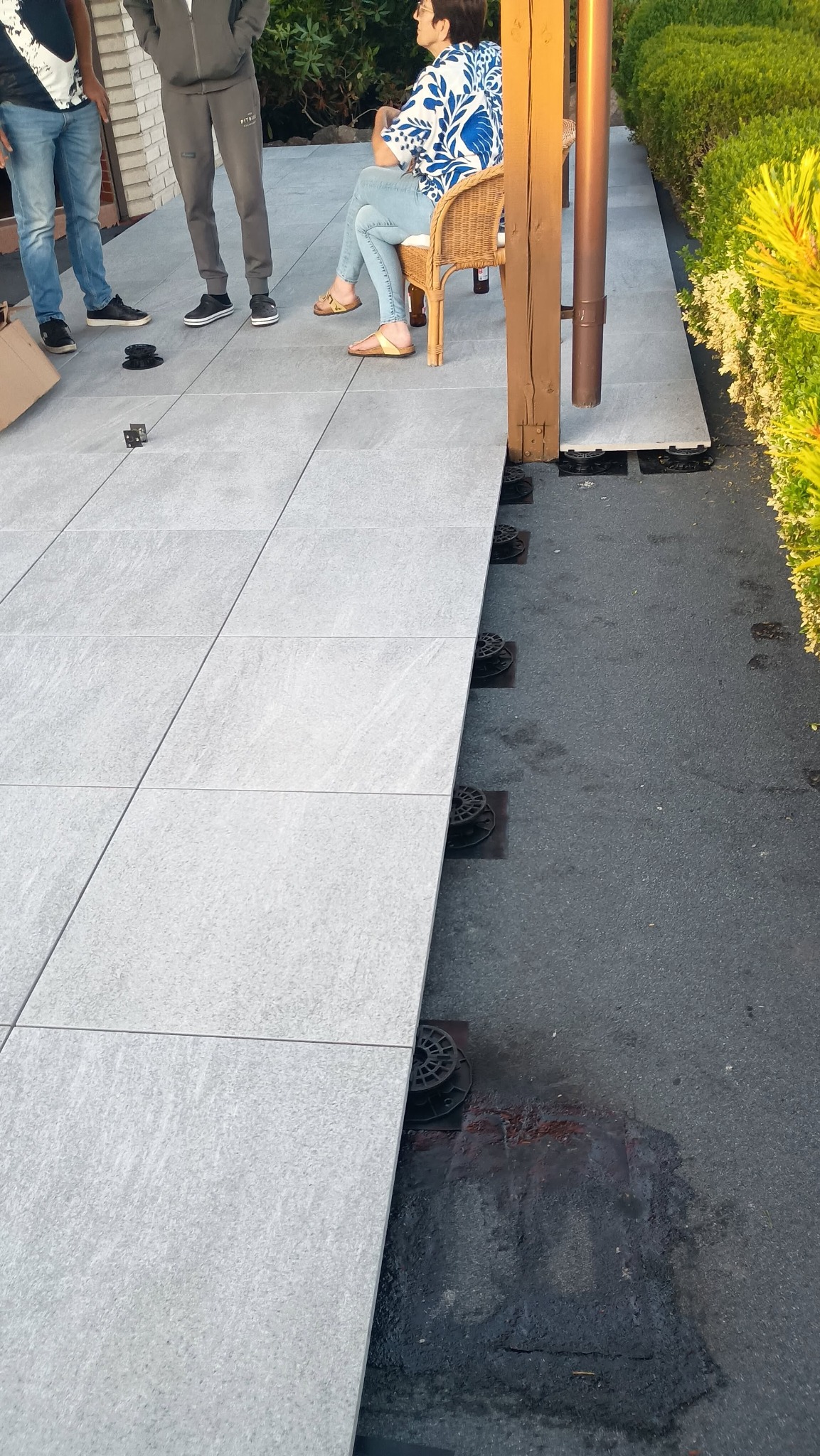 Montaż jasnoszarego tarasu z płyt na regulowanych wspornikach. Częściowo ułożone płyty, widoczne osoby i elementy konstrukcyjne. Ujęcie z góry, w dziennym świetle.