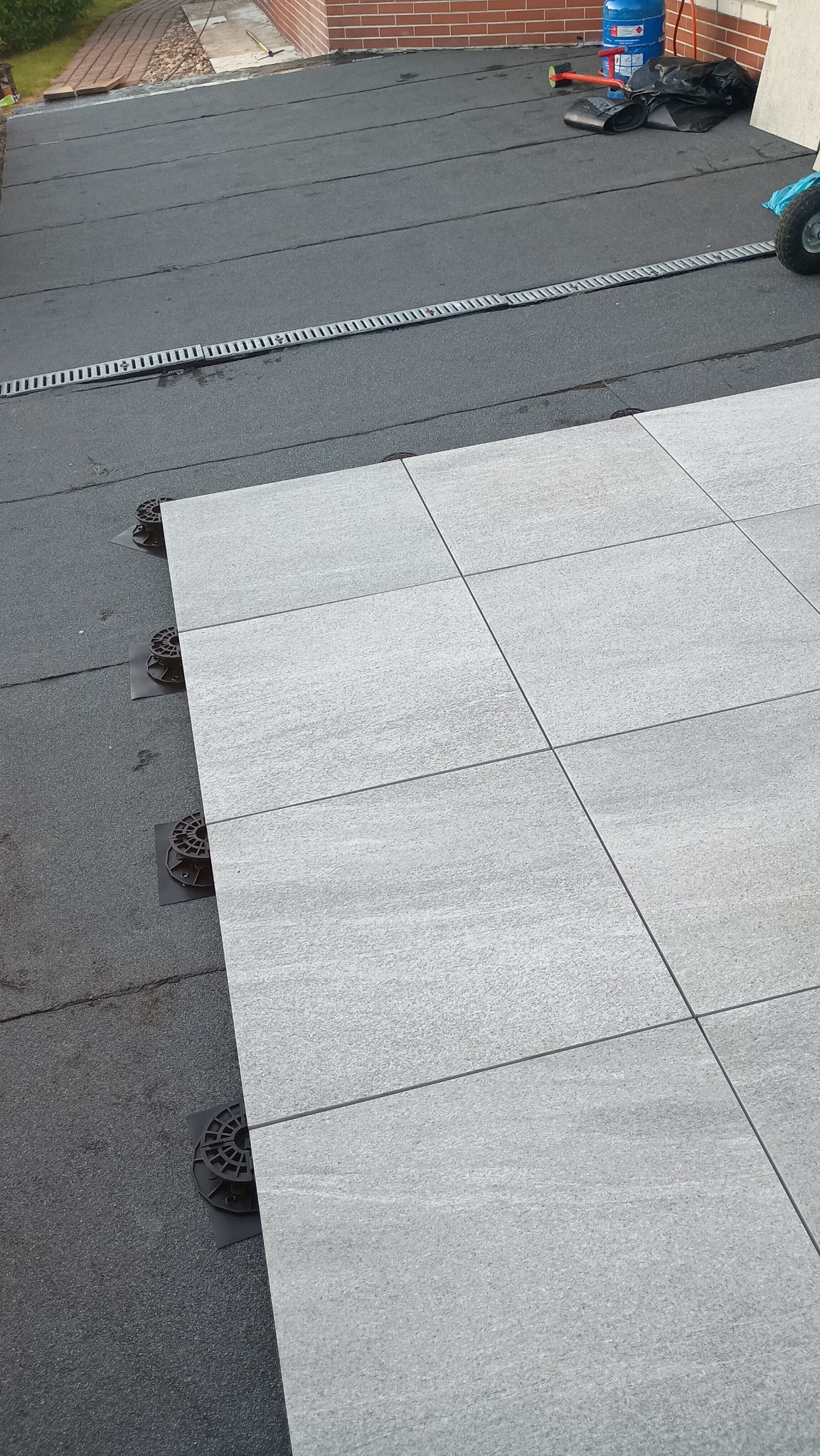 Montaż szarej, kwadratowej płyty tarasowej na dachu z papy, wspartej na regulowanych wspornikach. Widoczny fragment dachu z odpływem liniowym i narzędzia w tle.