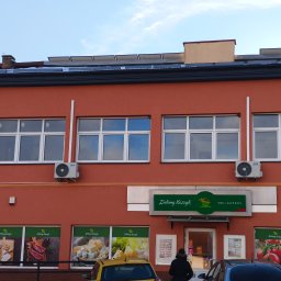 DG ALLINSTAL Dominik Goleń - Czerwony budynek sklepu z klimatyzacją AUX pod oknami. Na dachu panele słoneczne. Widoczne okna z białymi ramami i szyld sklepu spożywczego.